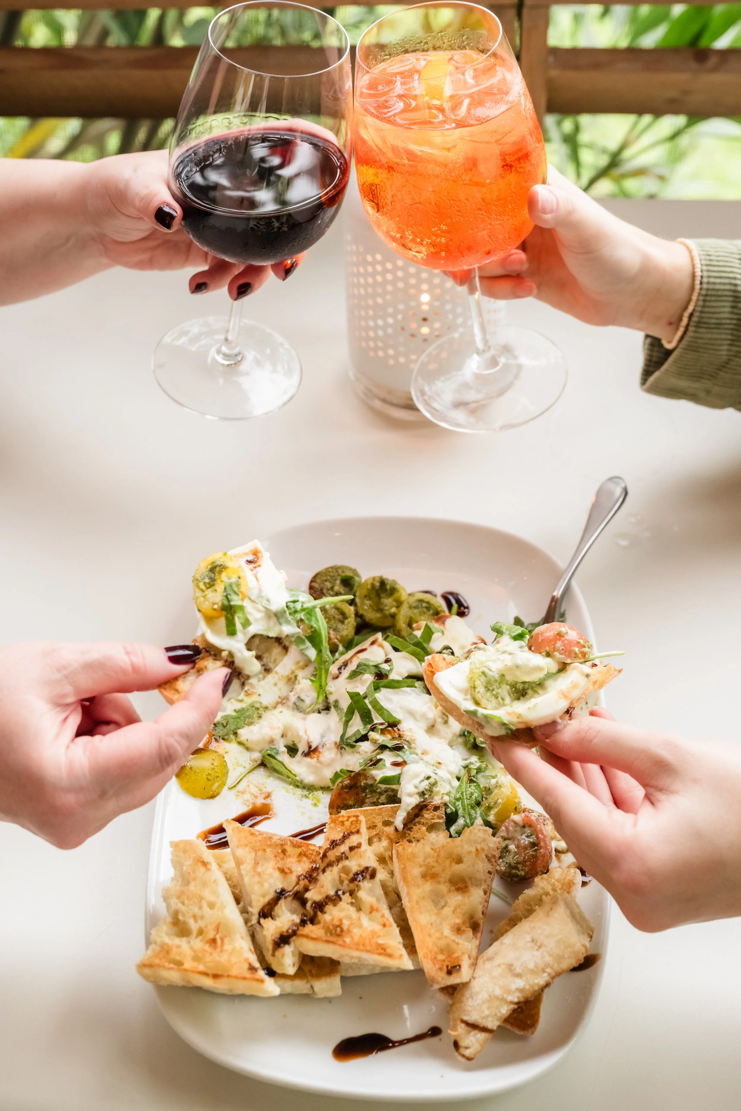 Two people clinking glasses of red and orange drinks over a plate of assorted appetizers including bruschetta with sliced bread and topped with cheese, herbs, and olives, on a white table with a candle in the background.