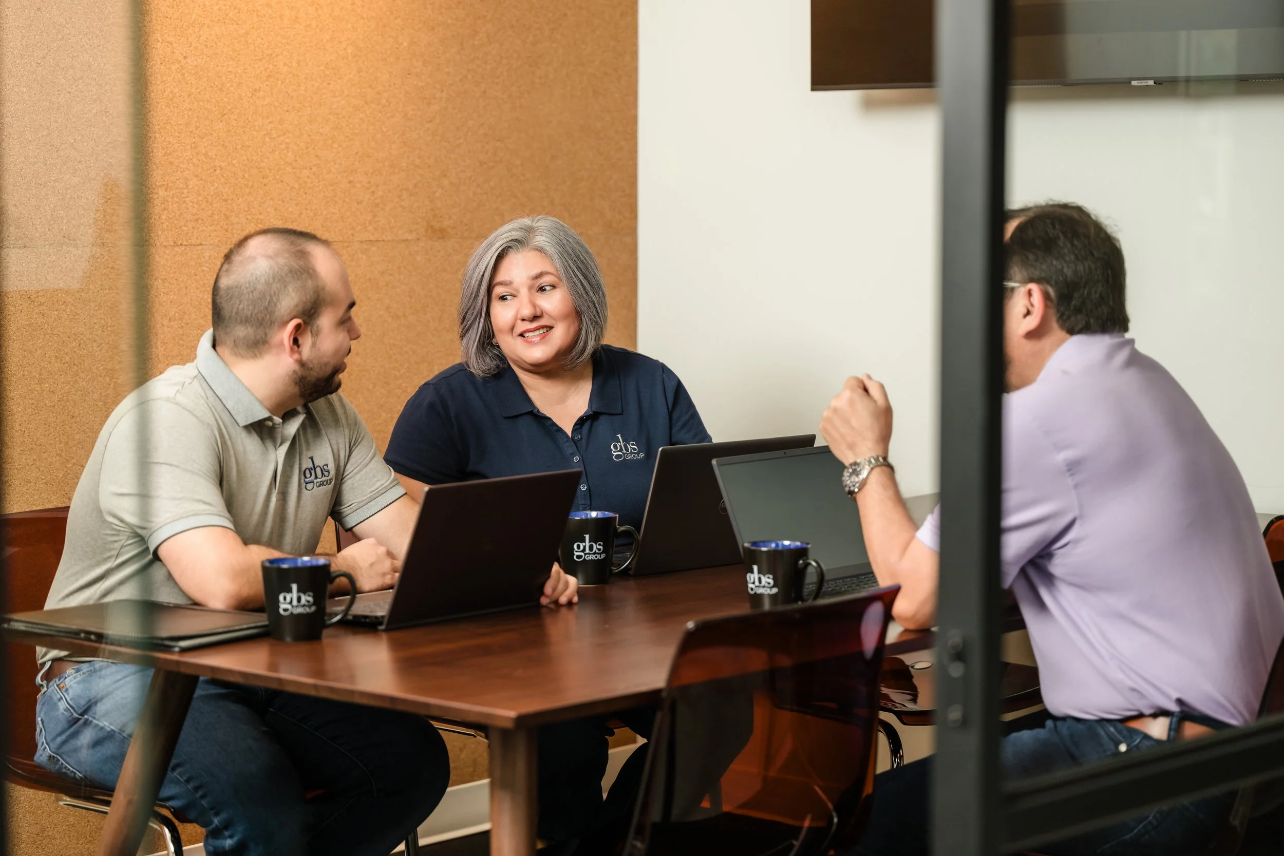 Four colleagues having a meeting in a conference room, with laptops and coffee mugs, engaged in conversation.
