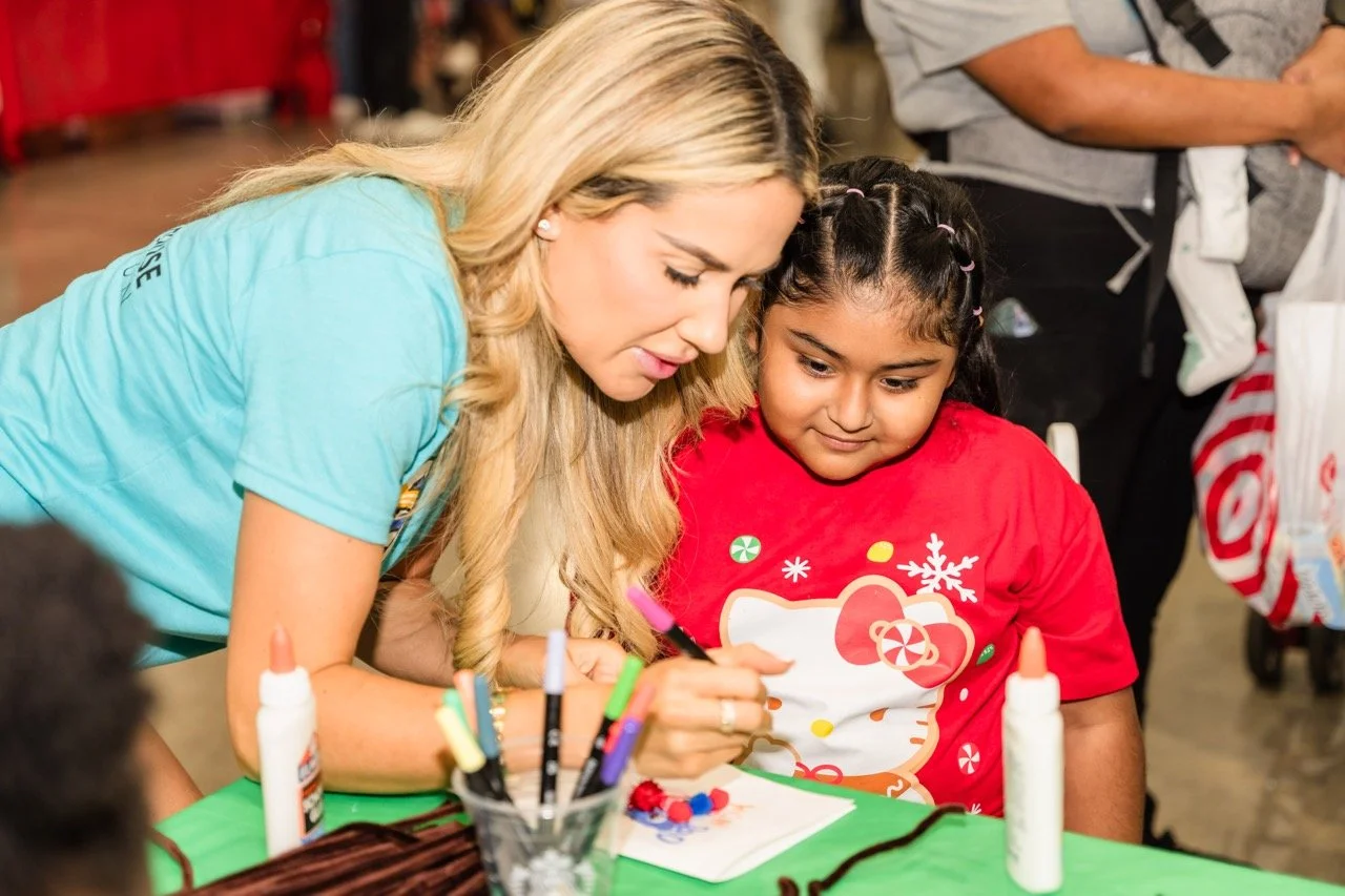 A woman helping a young girl with a holiday-themed craft activity at a table covered with green paper, decorated with colorful pens, glue, and yarn.