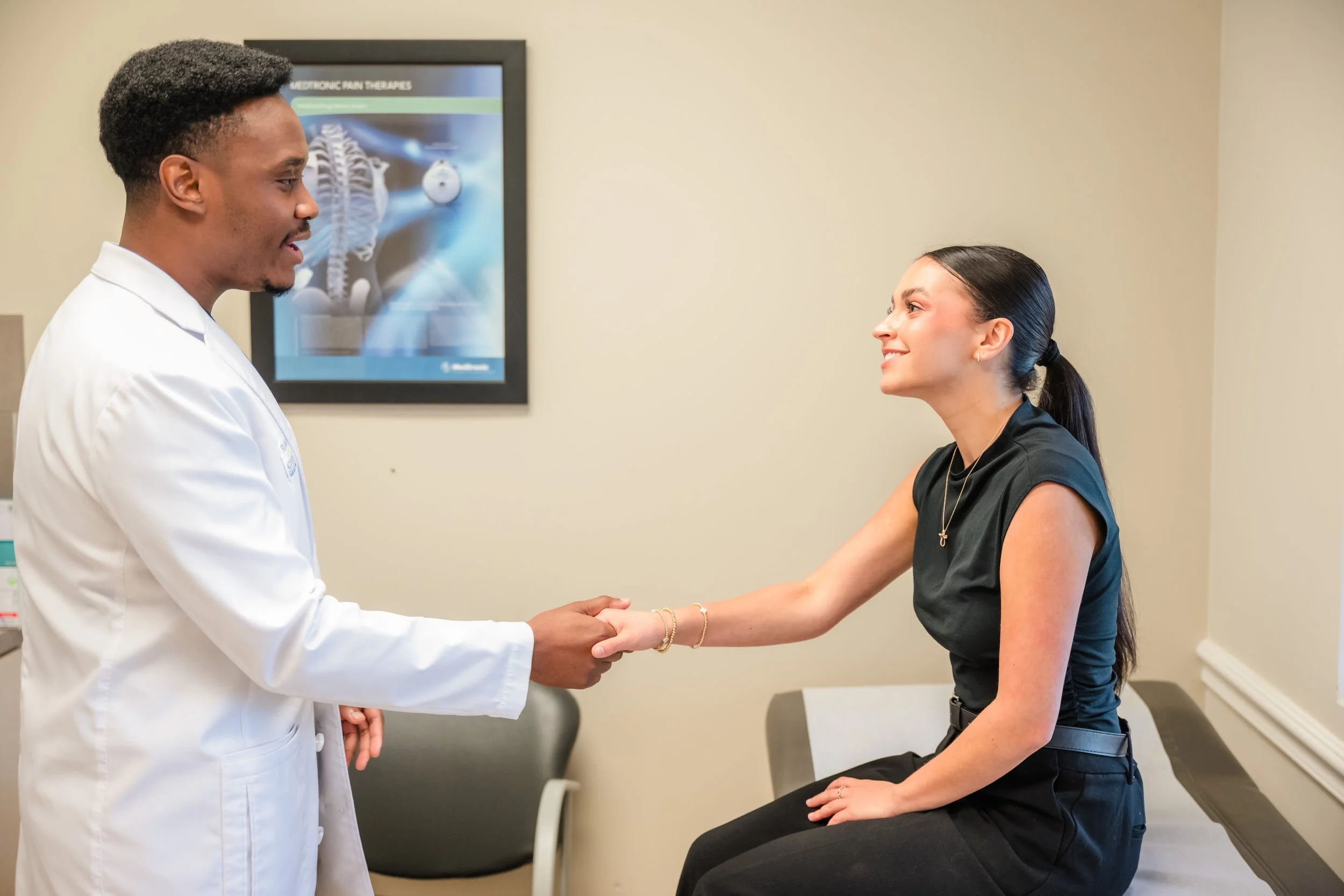 A male doctor in a white coat shaking hands with a female patient seated on an examination table in a medical office.