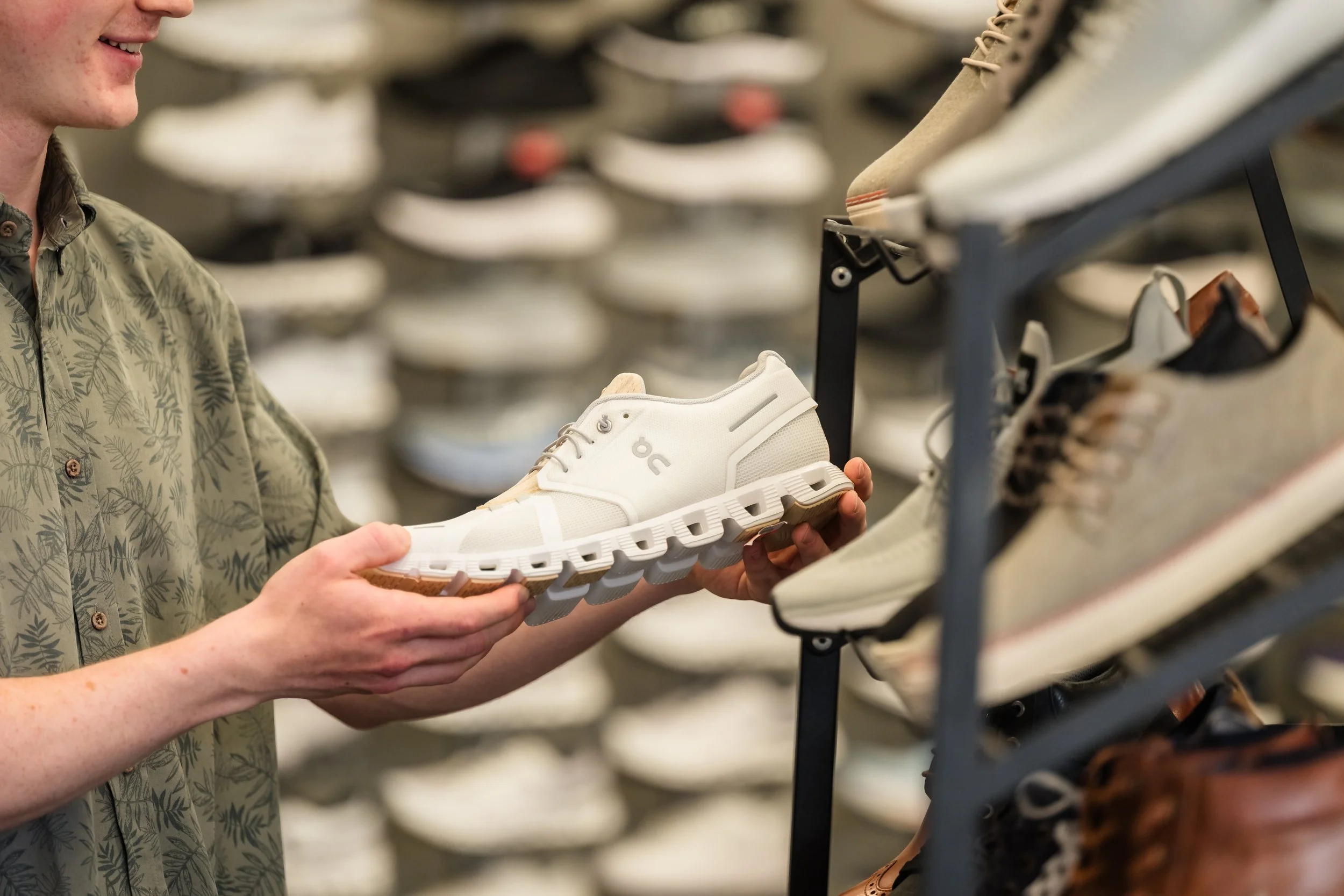 A person holding a white athletic shoe in a shoe store, surrounded by shelves with various sneakers.