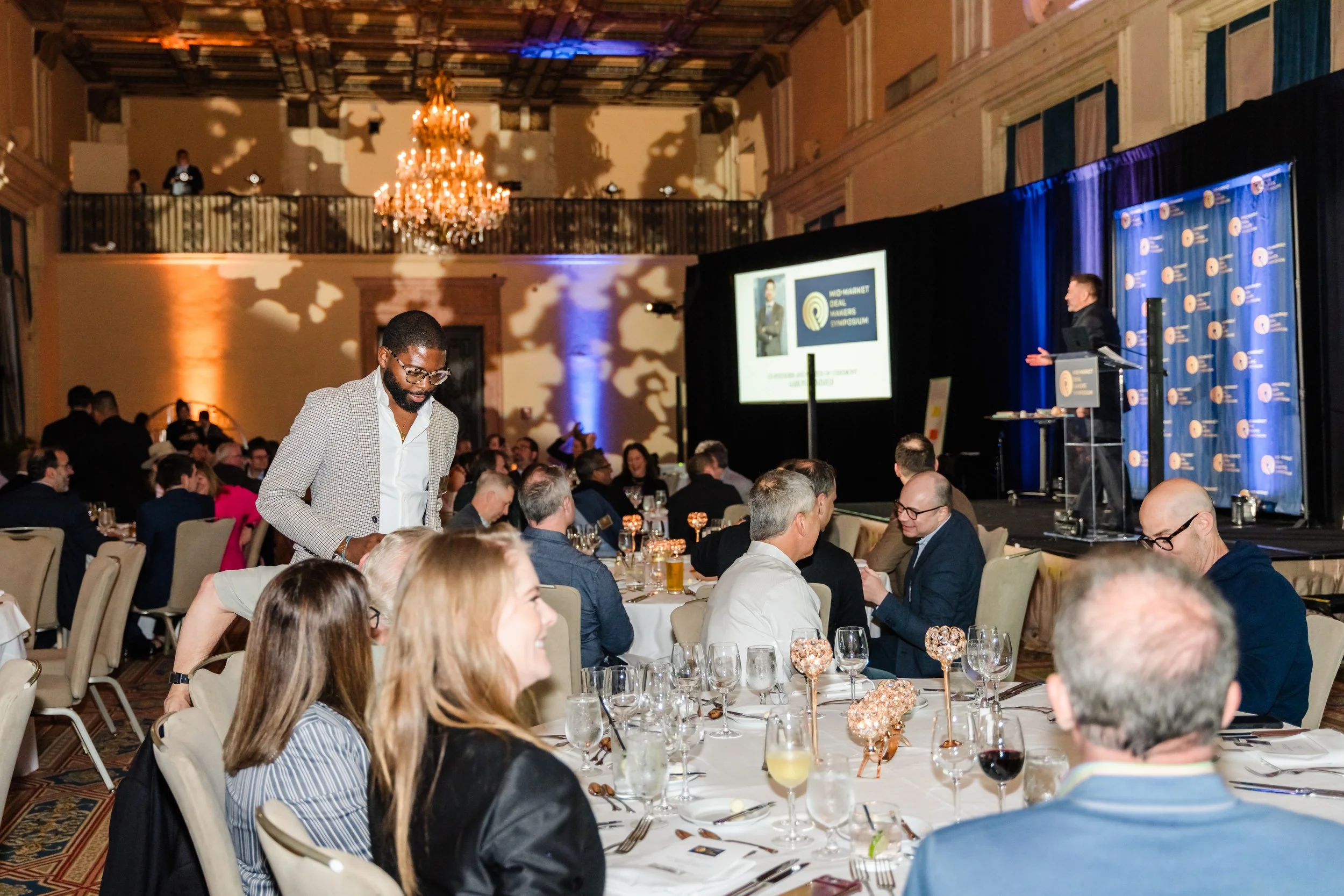 People attending a formal conference or gala in an ornate ballroom with a large chandelier, seated at round tables with drinks and decor, listening to a speaker on stage.