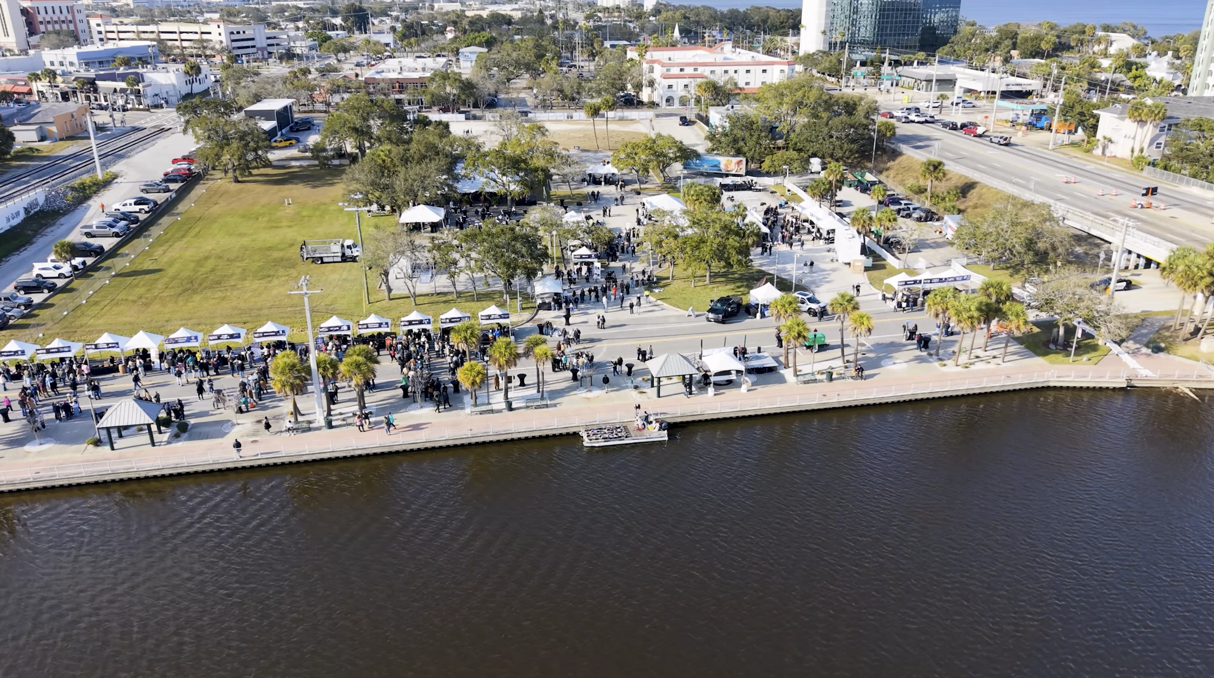 Aerial drone view of the Space Coast Food & Wine Festival along the waterfront riverwalk featuring white vendor tents, large crowds, palm trees, and downtown buildings on a sunny day in Florida.