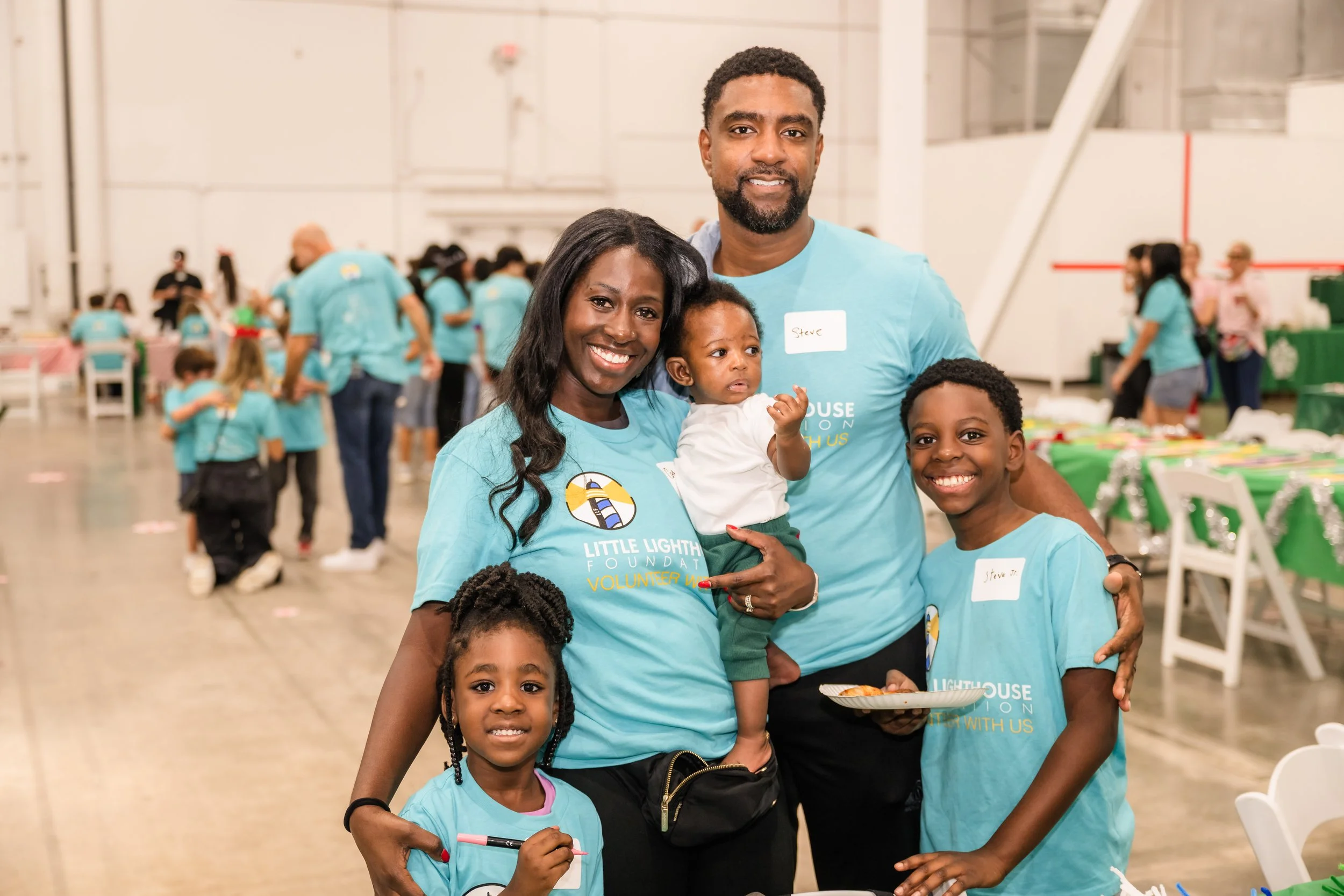 A family of five smiling indoors at a community event, wearing matching blue shirts with the Little Lighthouse Foundation logo, with children and other people in the background.