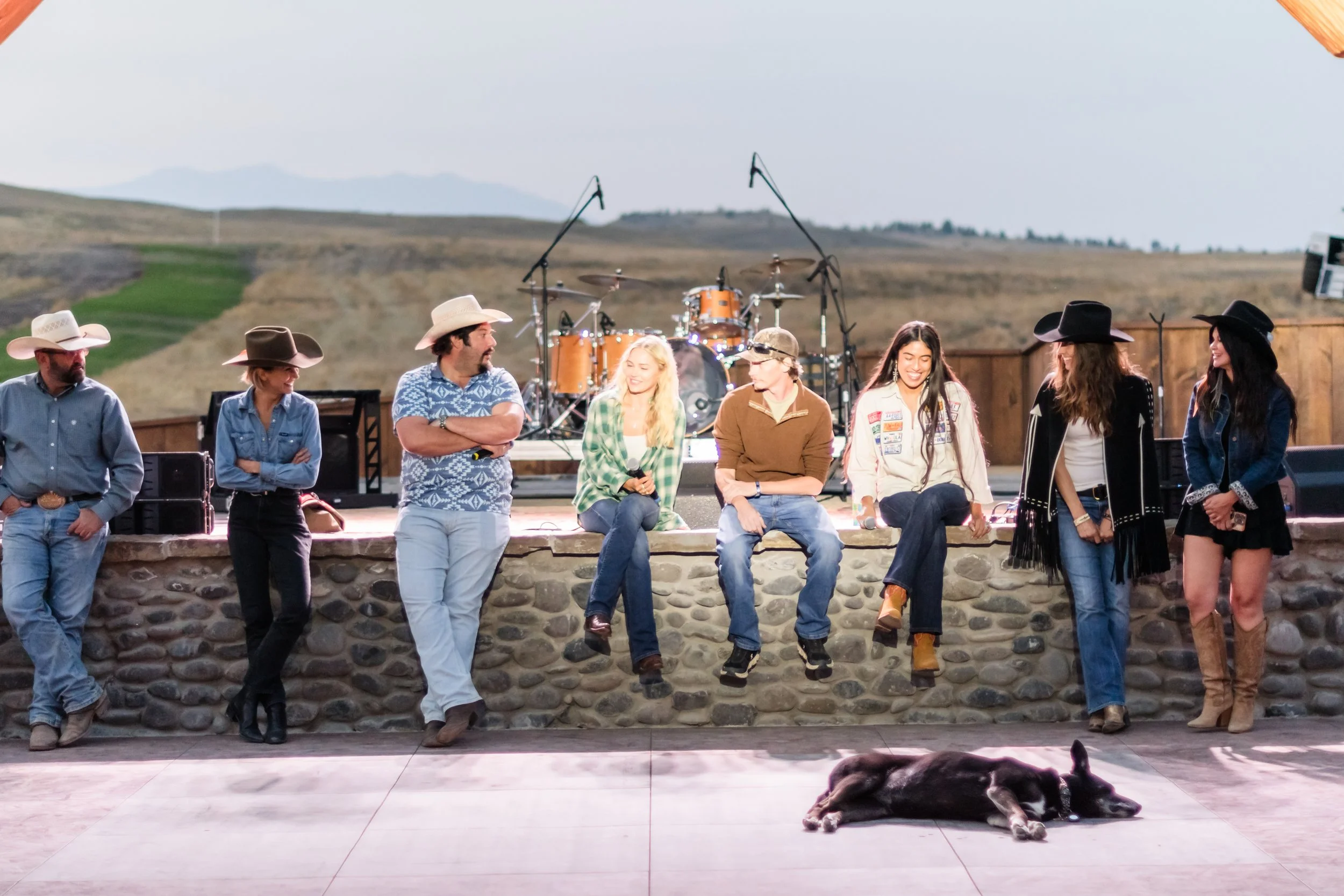 A group of people dressed in cowboy attire standing and sitting on a stone ledge in front of a stage with drums and microphones, outdoors with open fields in the background, and a black and white dog lying on the ground in the foreground.