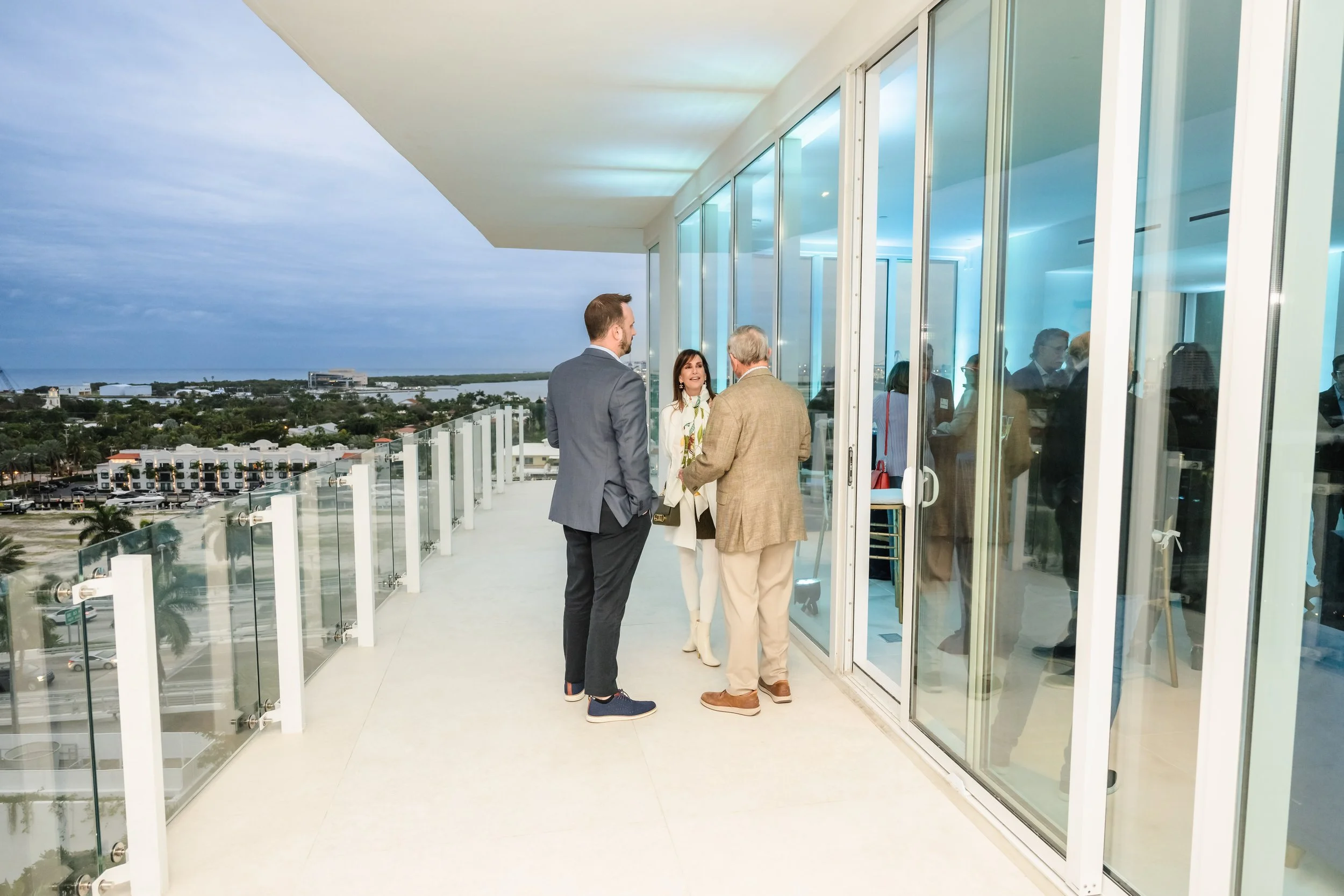 Three people engaged in conversation on a high-rise balcony with a city and water view.