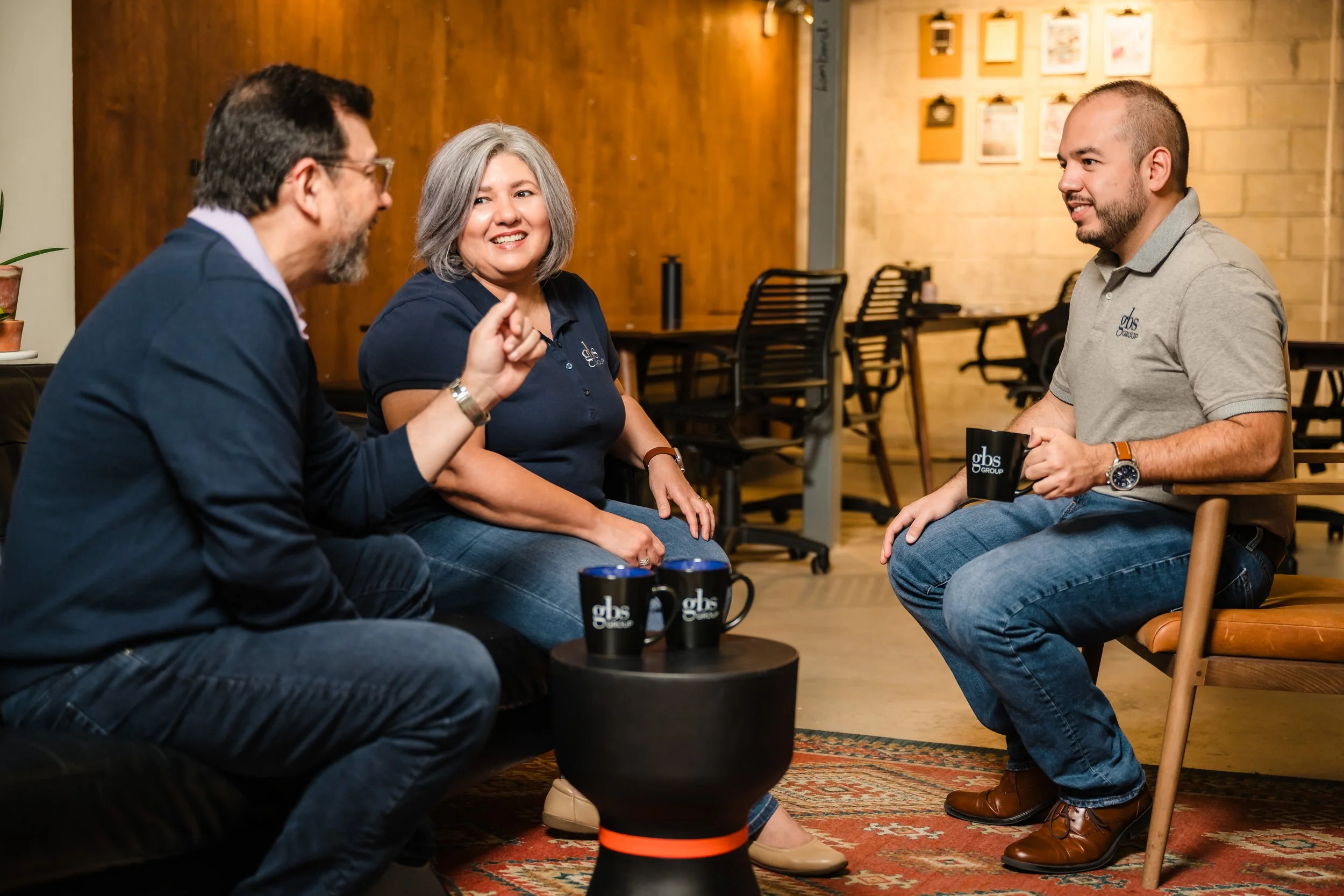 Three people sitting and talking in a casual office or workspace. They are holding black coffee mugs with the logo 'gbs' on them. The man on the left is gesturing with his hand, smiling, and wearing glasses, a dark blazer, and jeans. The woman in the
