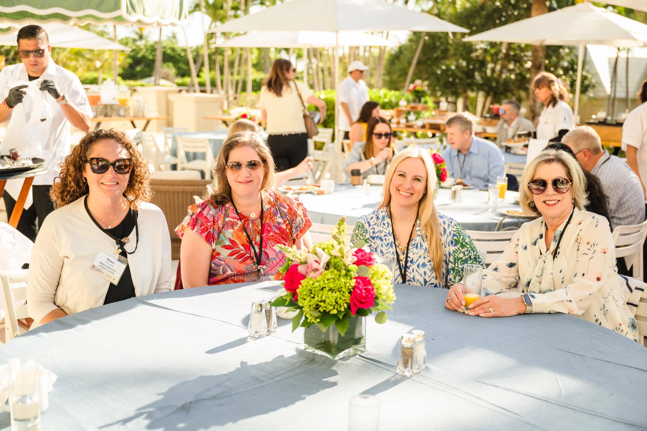 Four smiling women wearing sunglasses and conference lanyards pose together at a white linen-covered table with a pink and green floral centerpiece on a sunny tropical patio, with umbrellas, palm trees, and other diners in the background.