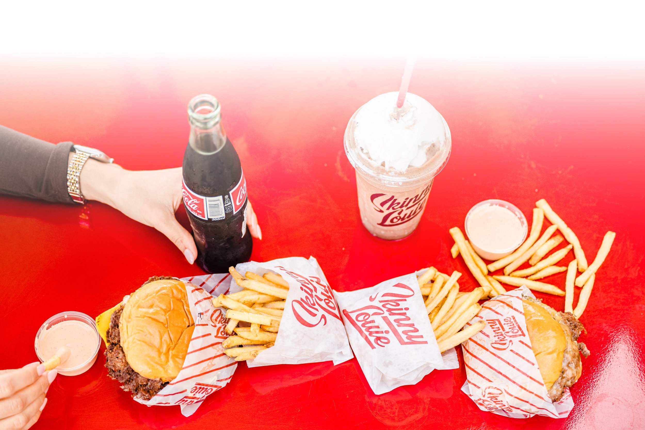 A meal at a red table featuring two cheeseburgers with buns, cheese, and meat patties, two portions of French fries, two small cups of dipping sauce, a bottle of Coca-Cola, and a mint Oreo milkshake with a straw.