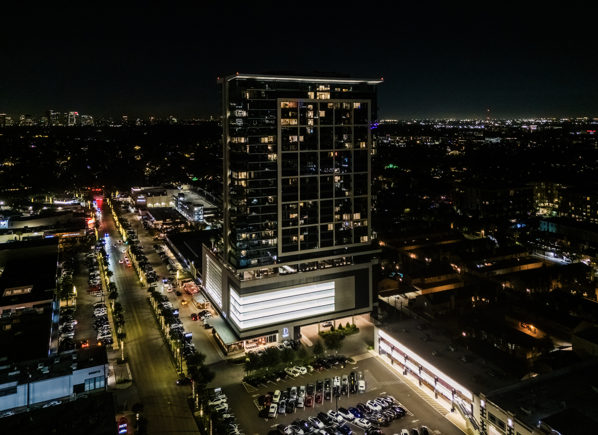 Nighttime cityscape with illuminated high-rise building, busy parking lot, and city skyline in the distance.