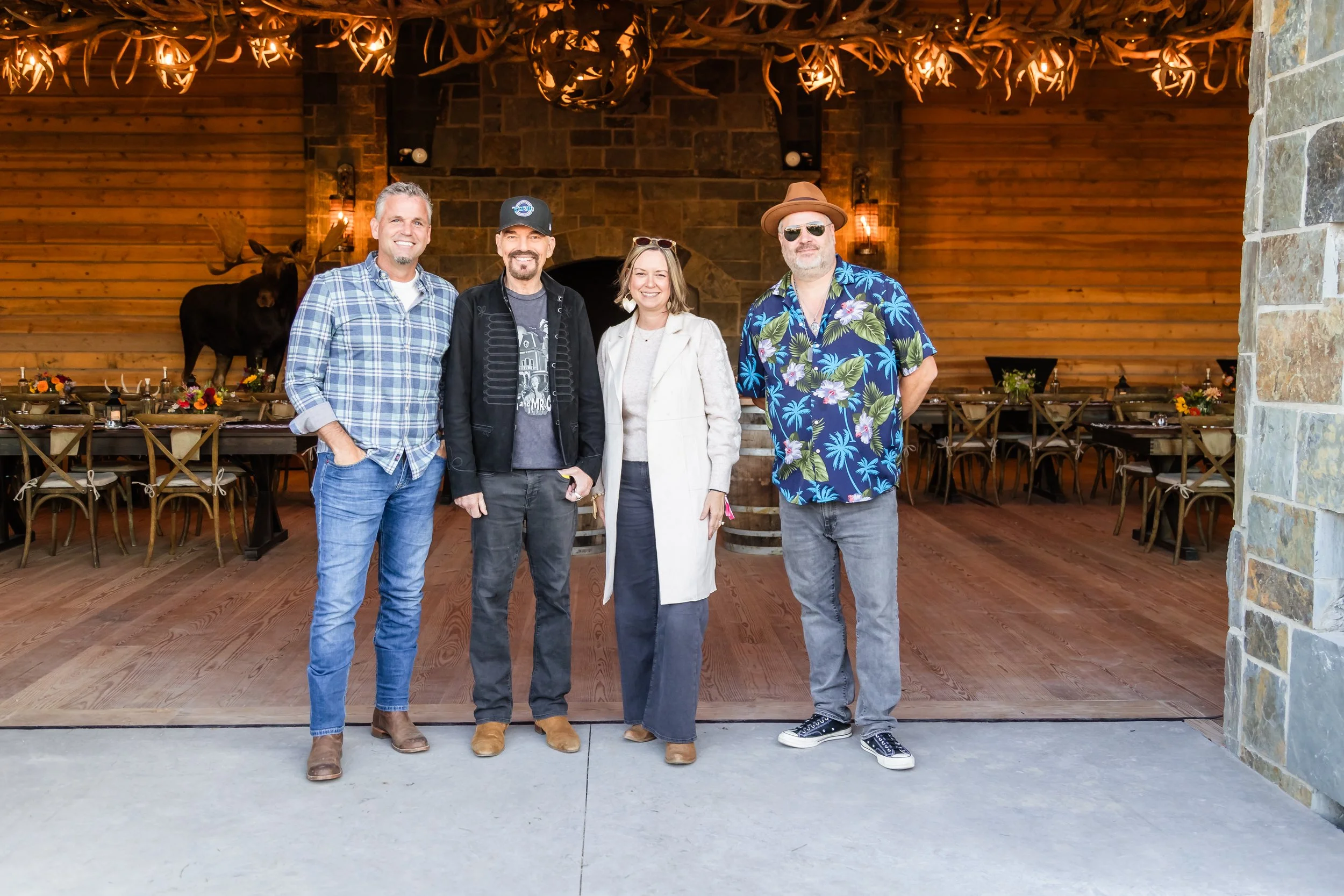 Billy Bob Thornton and J.D. Andrew of The Boxmasters posing with two guests during a meet and greet in front of a large stone fireplace with antler chandelier and warm lighting inside a rustic Montana lodge at the 2025 Bourbon and Bonfire event.