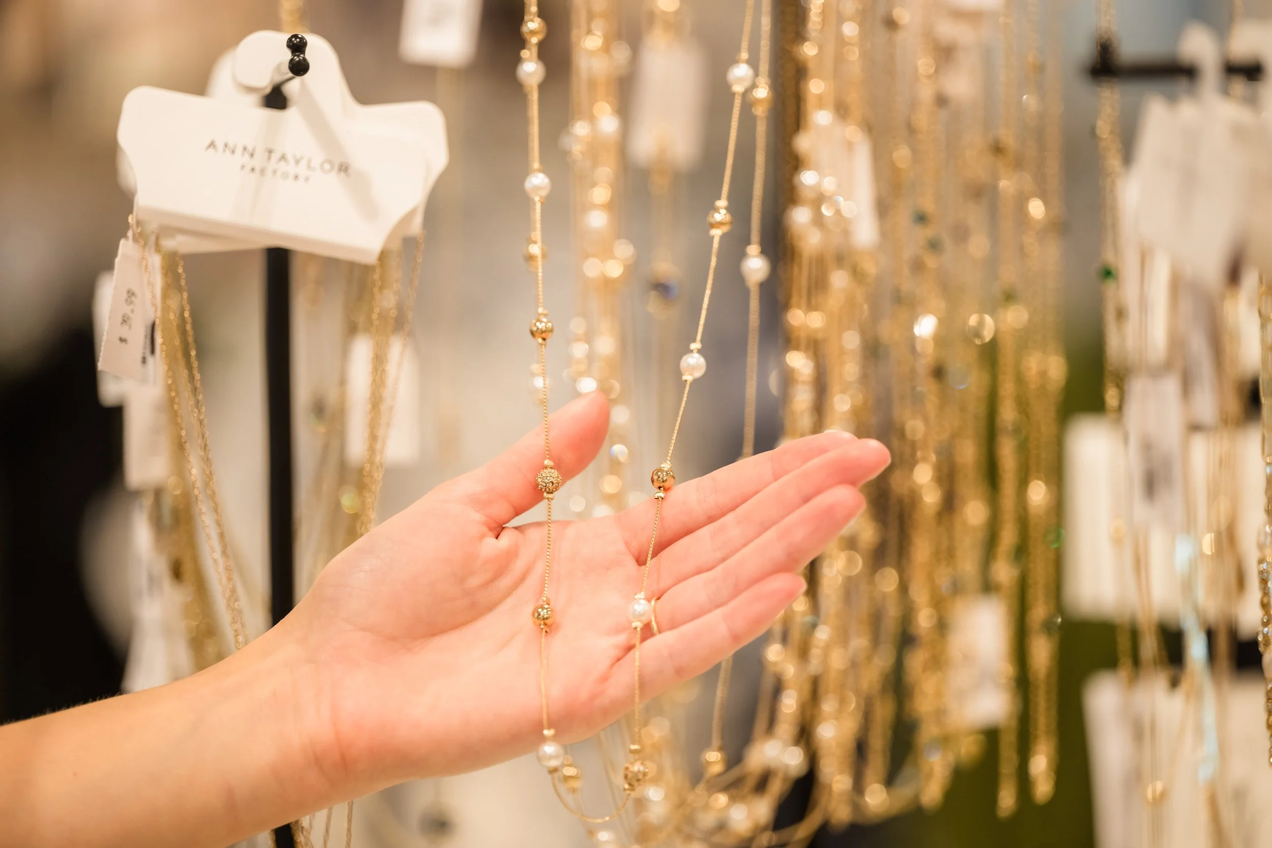 Close-up of a person's hand holding a delicate necklace with gold beads and pearls, with jewelry and necklaces hanging in the background at a jewelry store.