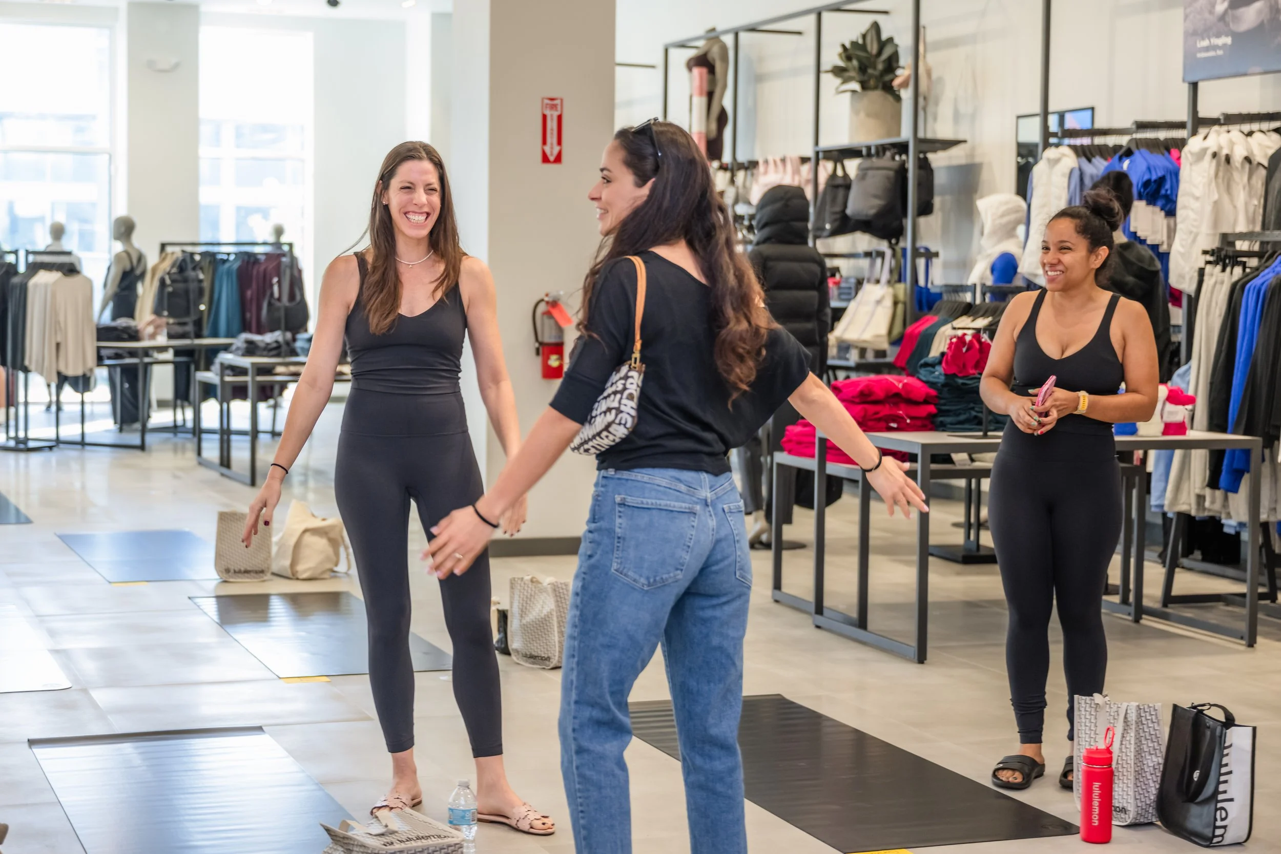 Three women inside a clothing store, two are engaging in a conversation, while the third stands nearby, smiling. The store has racks with clothes, and shopping bags on the floor.