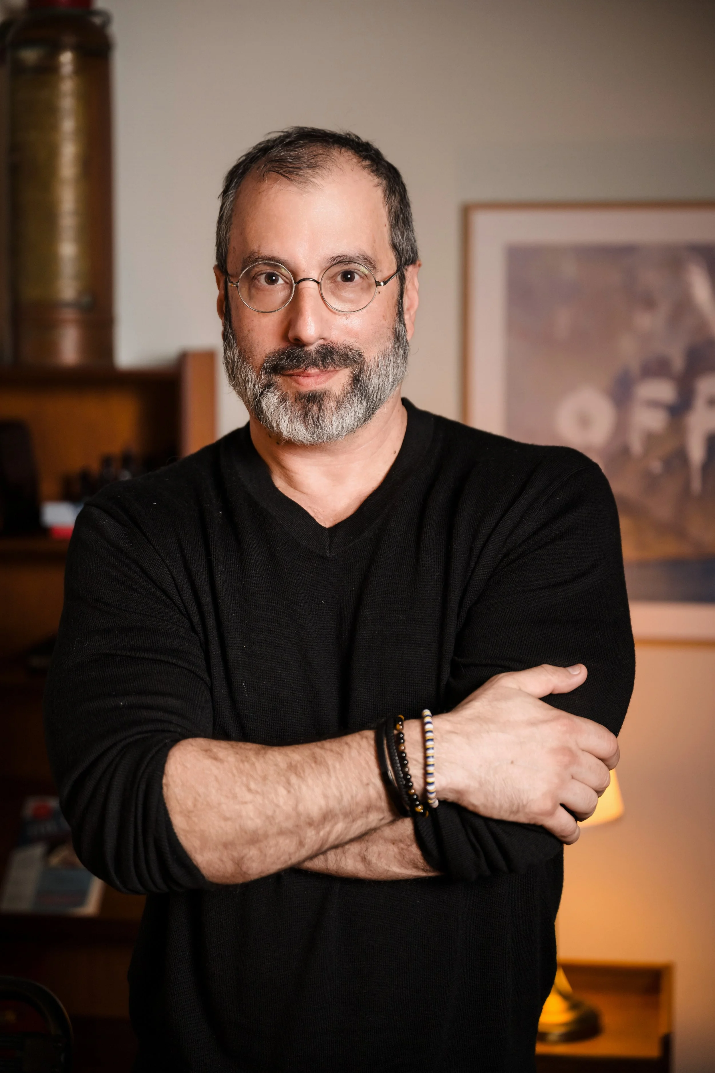 A middle-aged man with glasses and a gray beard, wearing a black sweater, crosses his arms and looks at the camera in a warmly lit room.