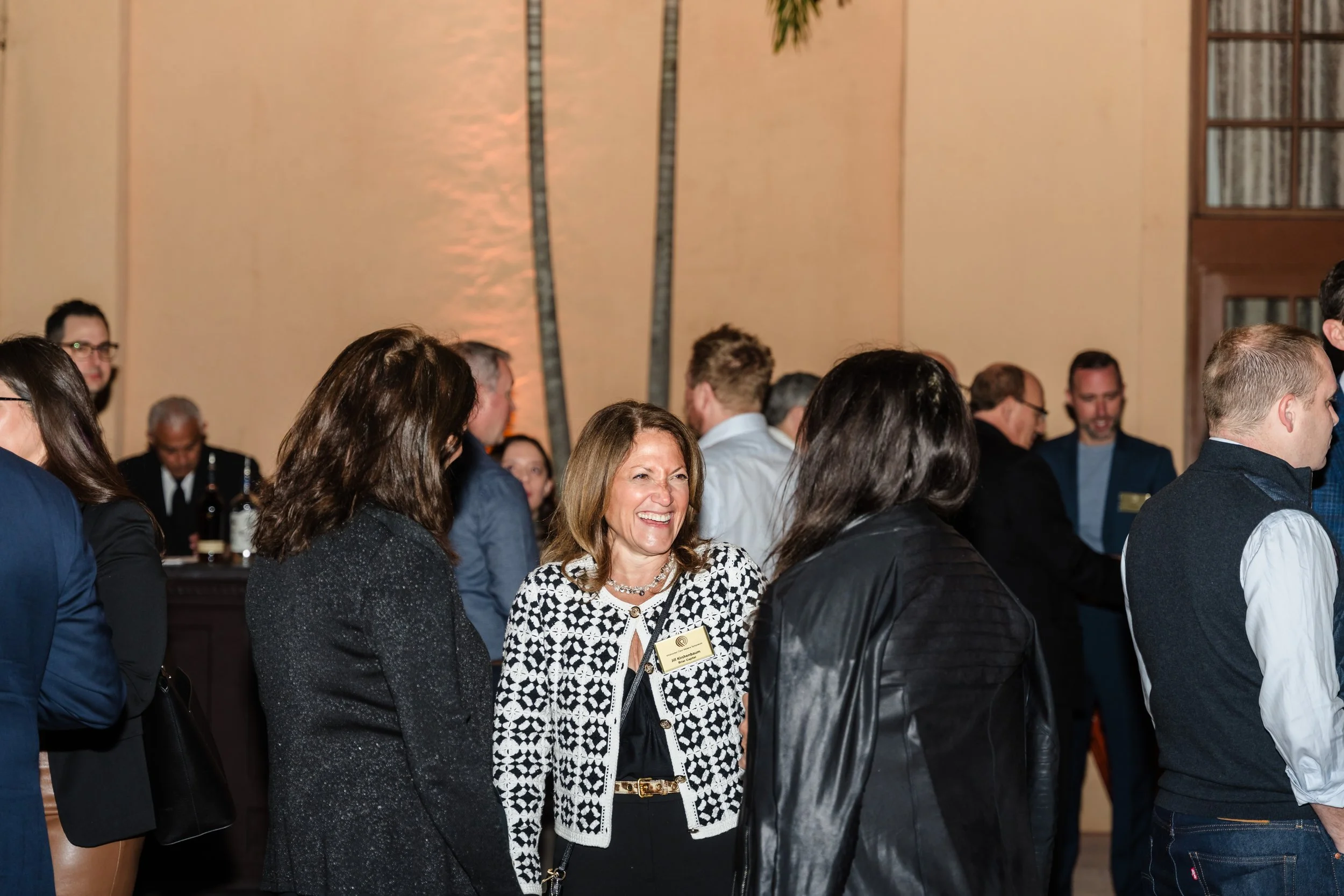 A woman in a black-and-white patterned jacket and event name badge smiles while chatting with fellow attendees during an indoor networking reception at The Biltmore, with palm trees and warm ambient lighting visible in the background.