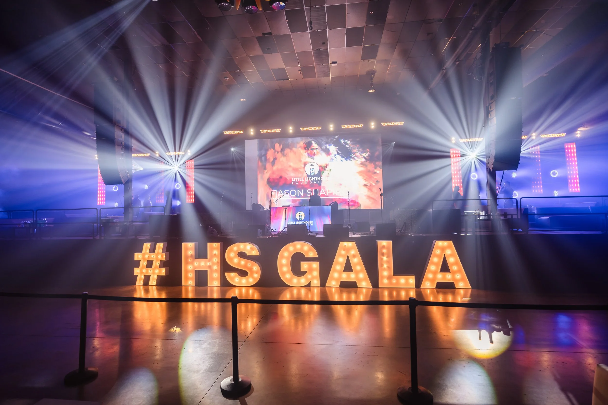 Stage set for a gala event with colorful lighting, large illuminated hashtag #HSGALA, a big screen displaying a logo and image, and a DJ booth in the background.