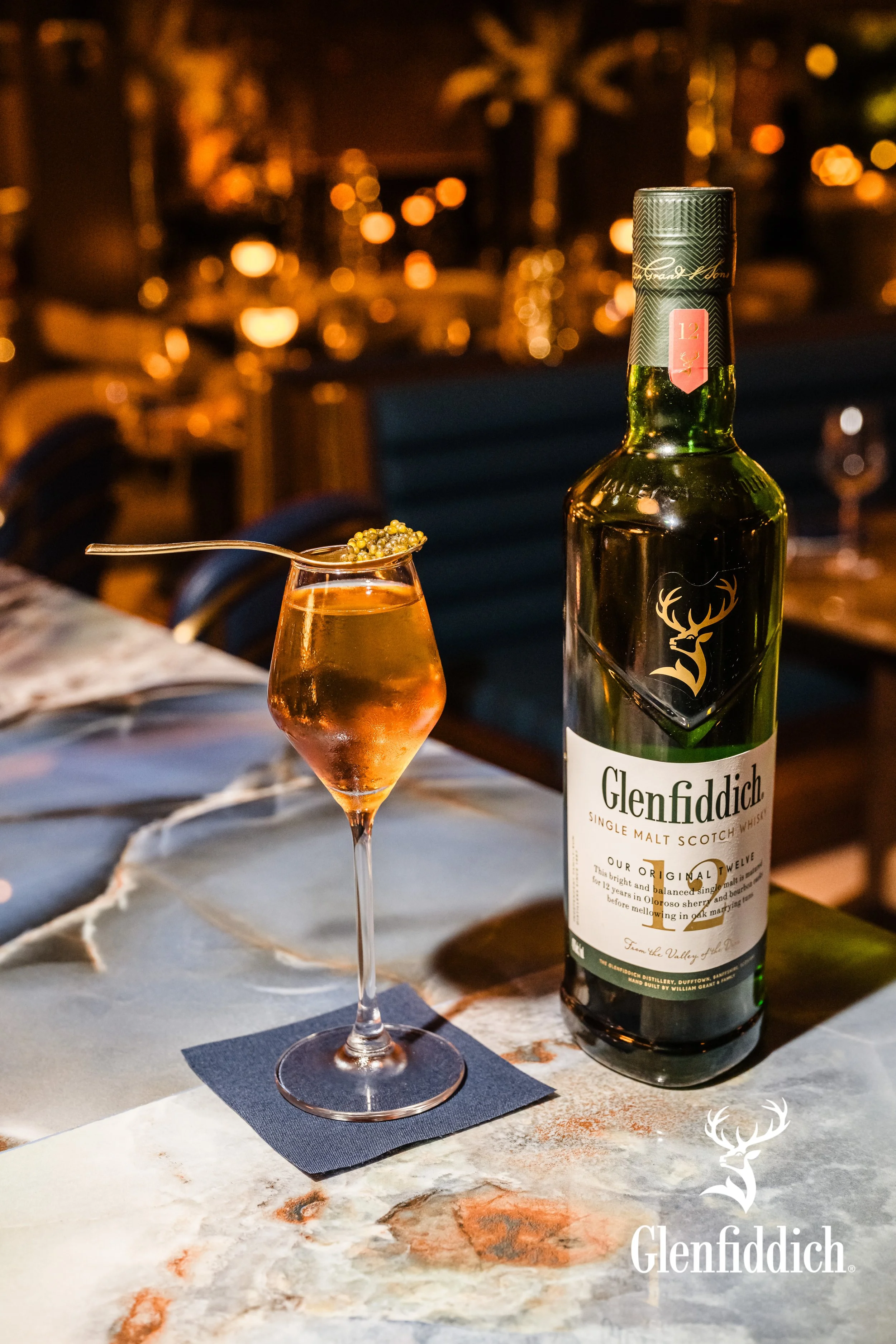 A bottle of Glenfiddich single malt scotch whiskey and a glass of whiskey with a garnish of greenish-yellow berries, on a marble table in a warmly lit restaurant or bar setting.