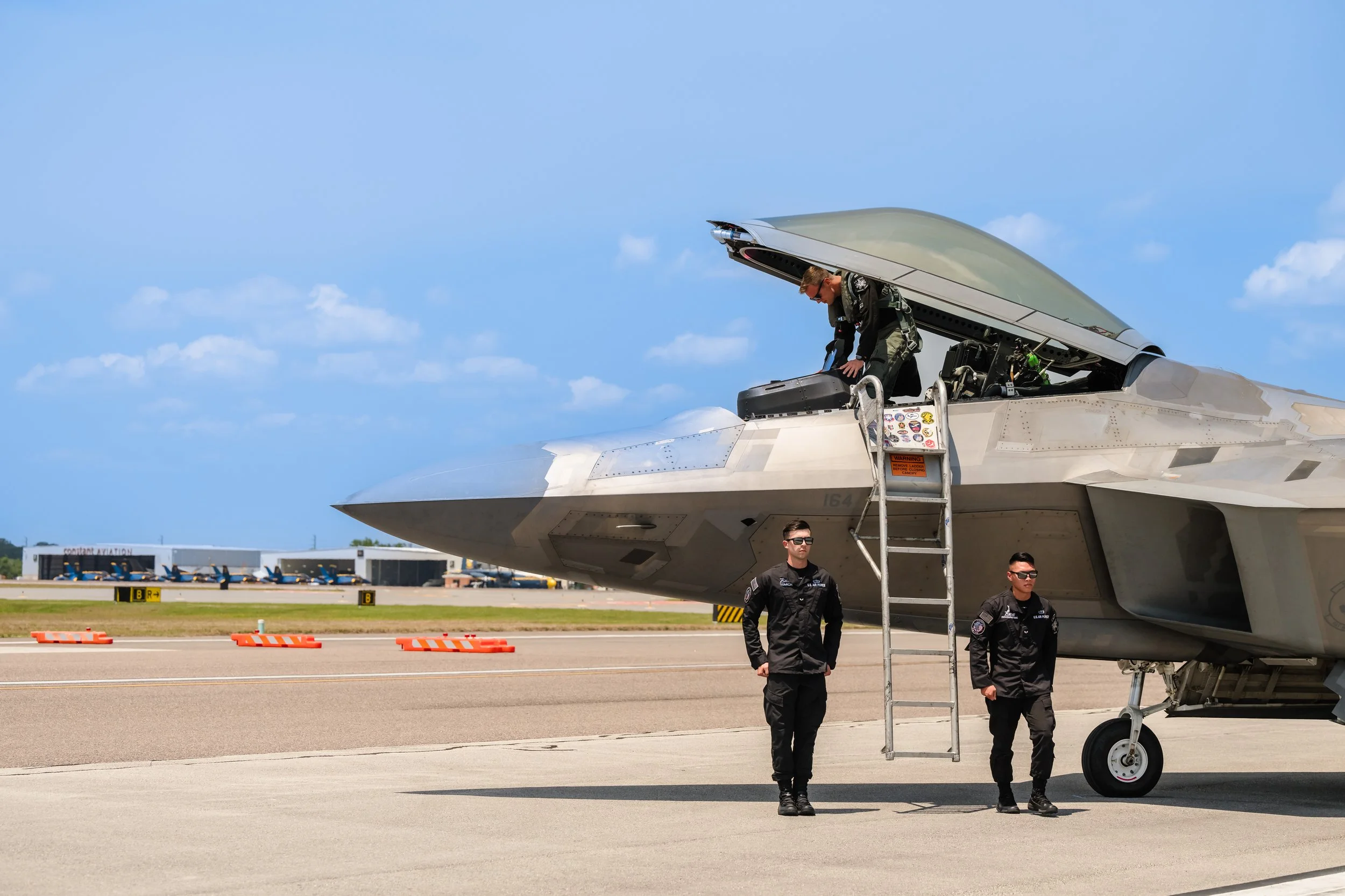 A fighter jet on an airstrip with one pilot leaning into the cockpit and two ground crew members walking nearby under a clear blue sky.