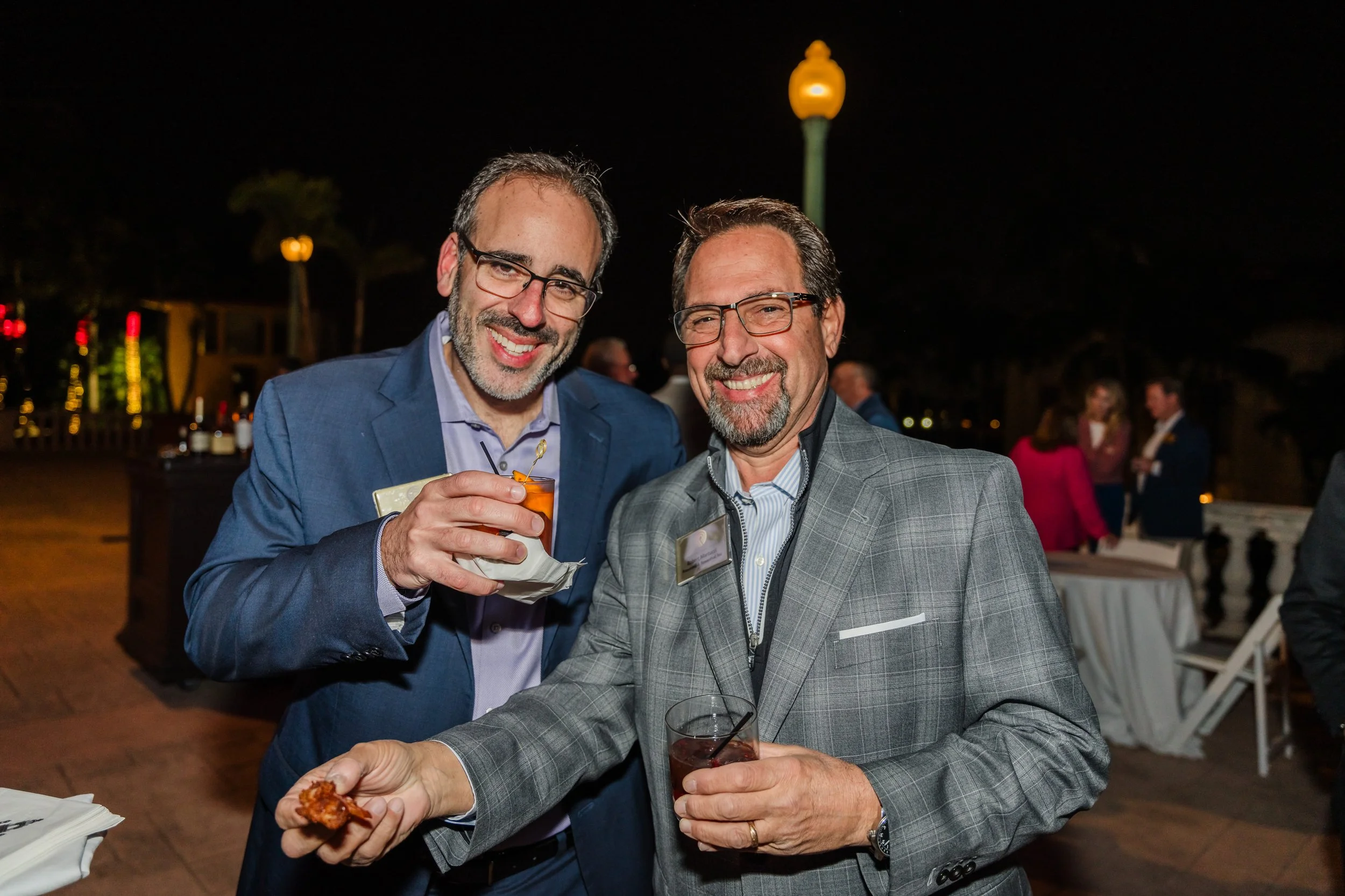 Two men in blazers smile for the camera while holding cocktails at an outdoor nighttime reception at The Biltmore during the Mid-Market Deal Maker Symposium 2025, with other guests and warm patio lighting in the background.