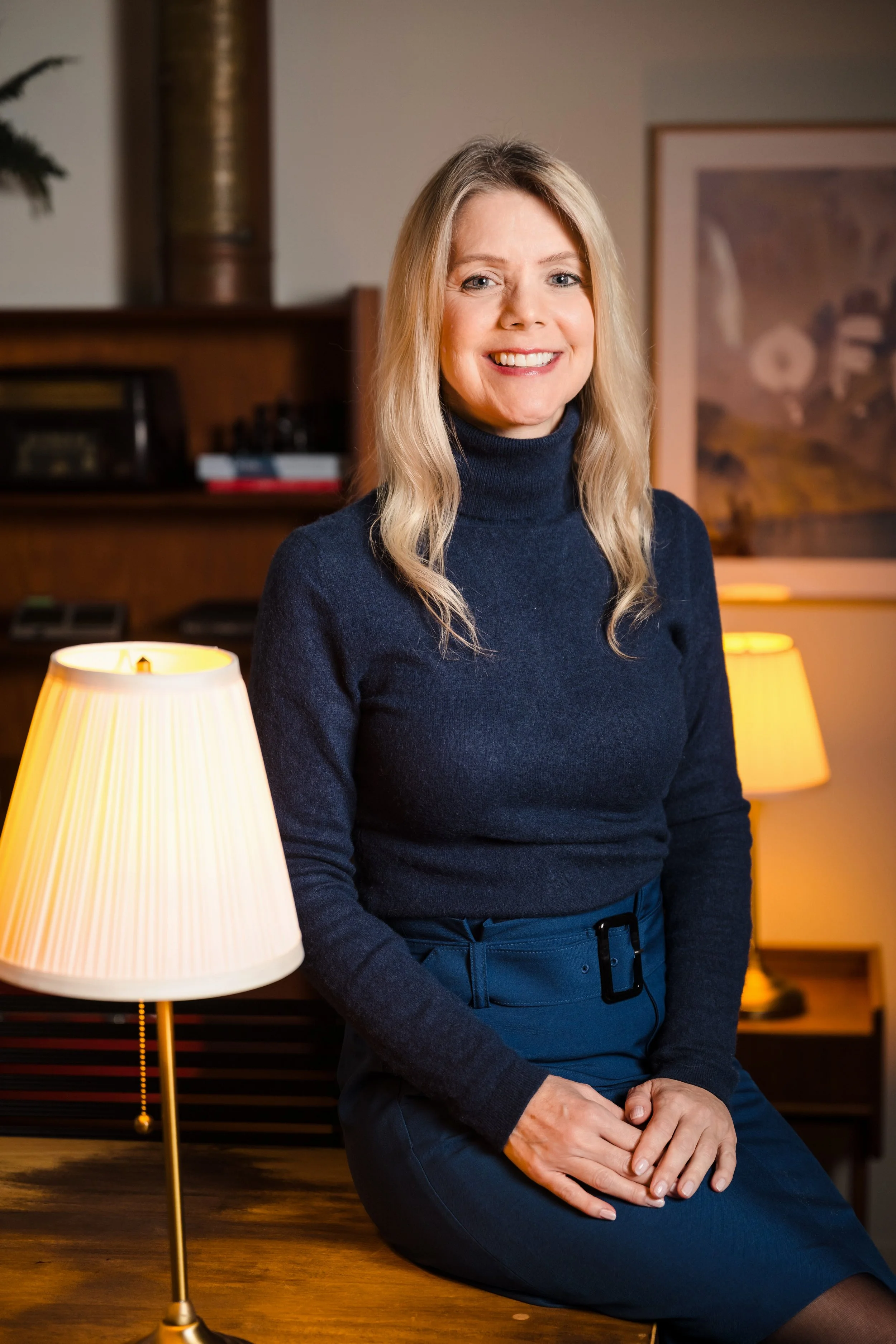 A woman with blonde hair wearing a navy turtleneck and dark skirt sitting on a wooden table in a warmly lit room.