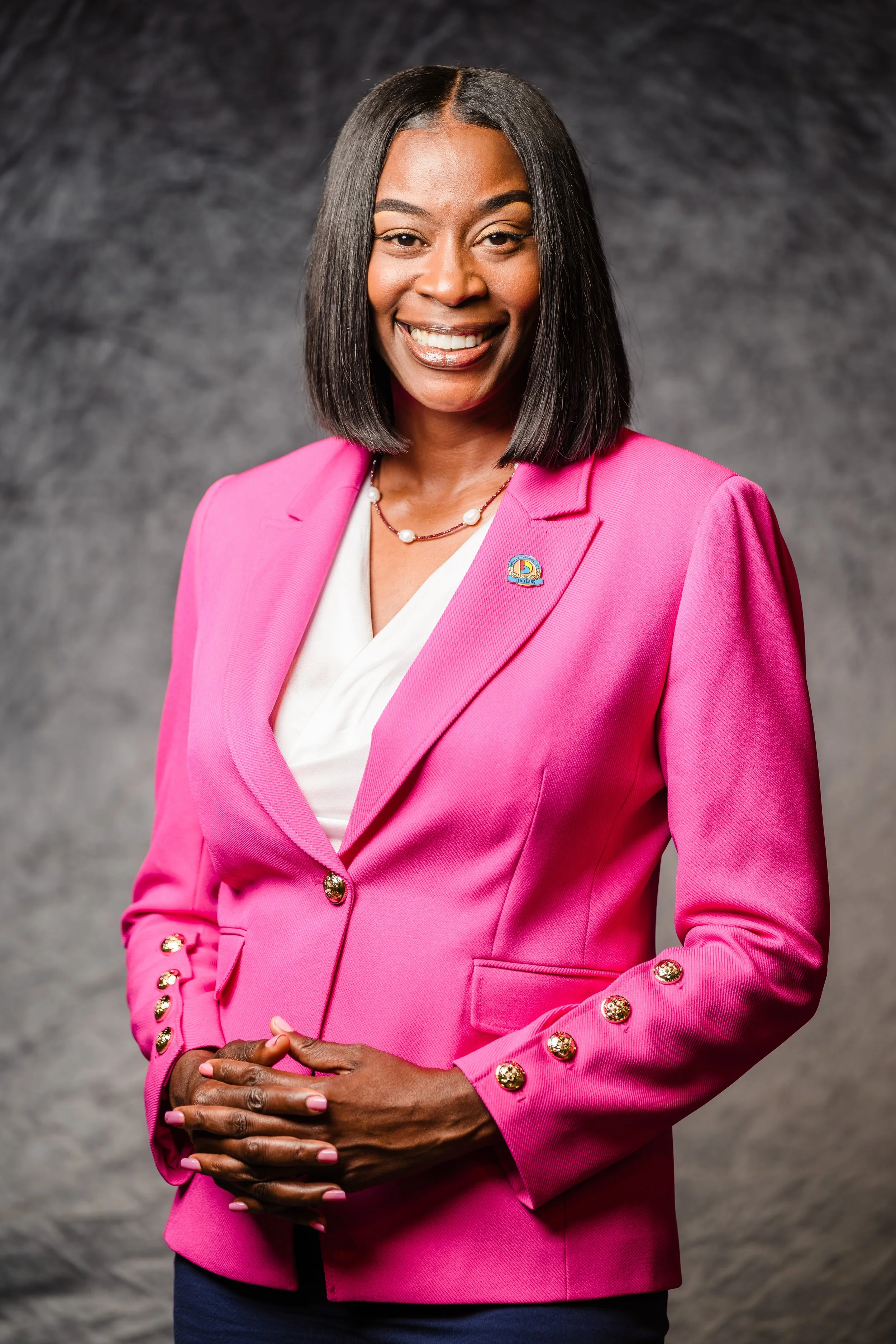 A woman with shoulder-length black hair wearing a bright pink blazer with gold buttons and a white top. She has a pearl necklace and a lapel pin, and is smiling with her hands clasped together in front of her. The background is a textured gray.