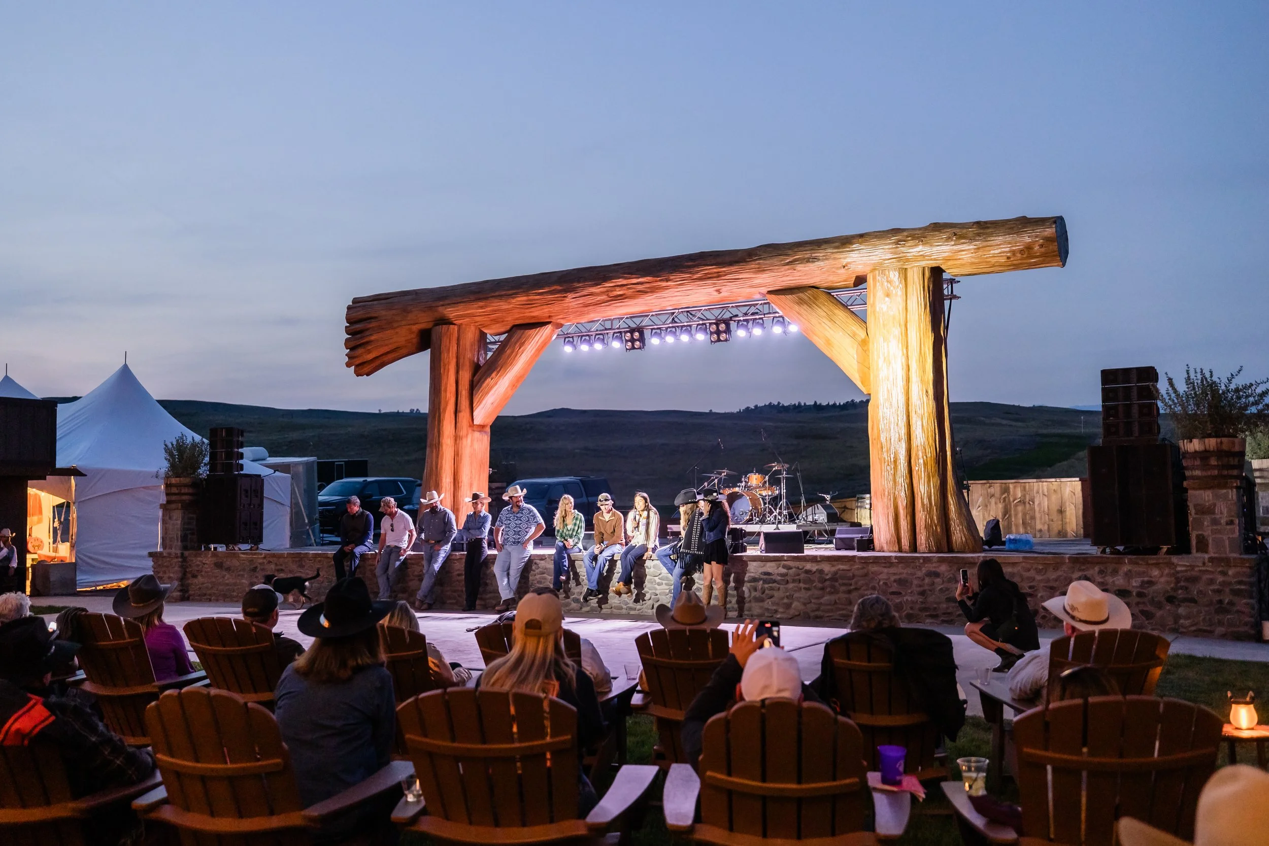 Outdoor concert stage with a large wooden arch and band members performing during dusk, audience seated on wooden chairs, some people taking photos, tents in the background, rolling hills under a cloudy sky.