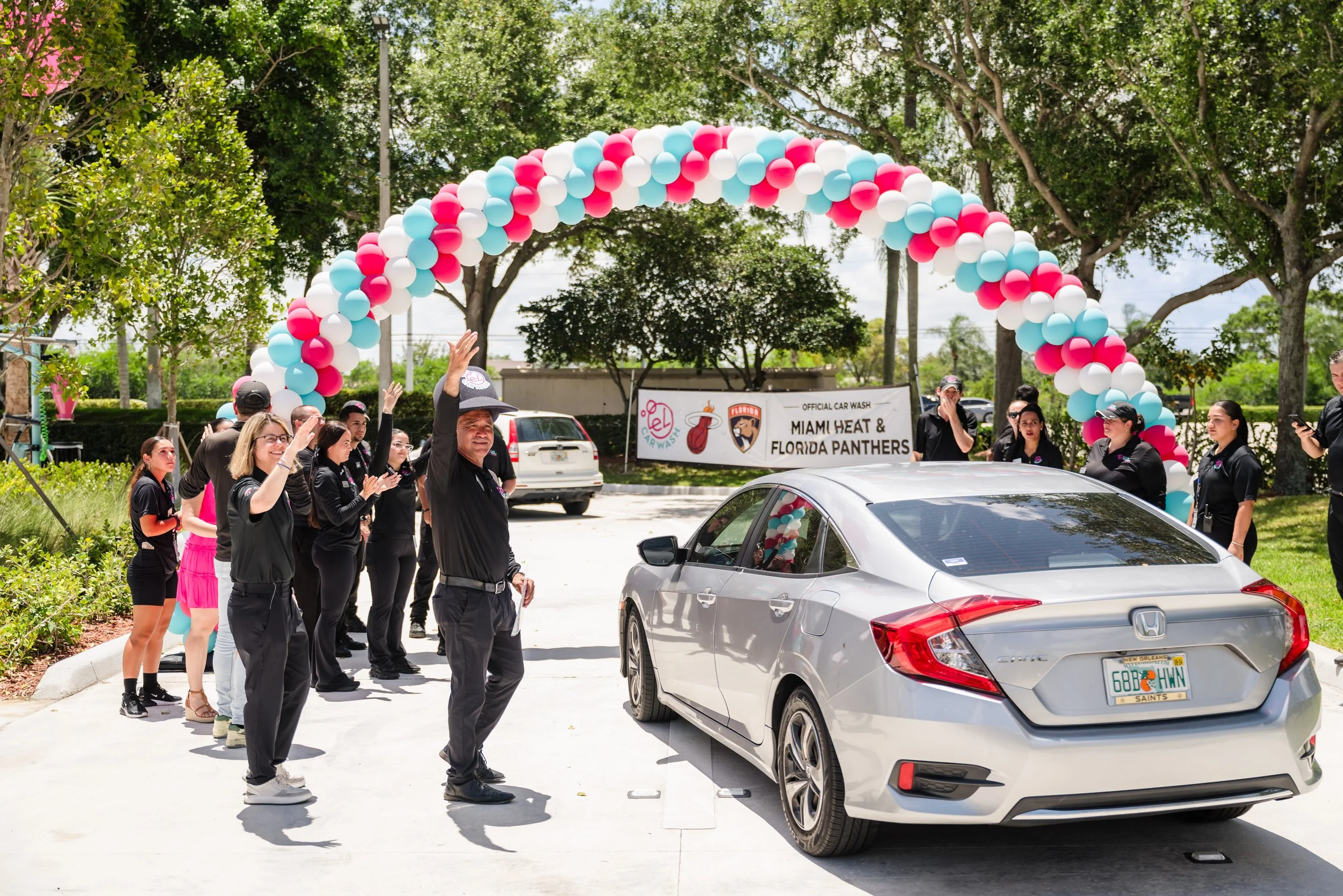 People gathered outside under a balloon arch, waving and clapping next to a silver Honda Civic with a Florida license plate, in a parking lot with trees and a sign that reads 'Official Car Wash Miami Heat & Florida Panthers'.