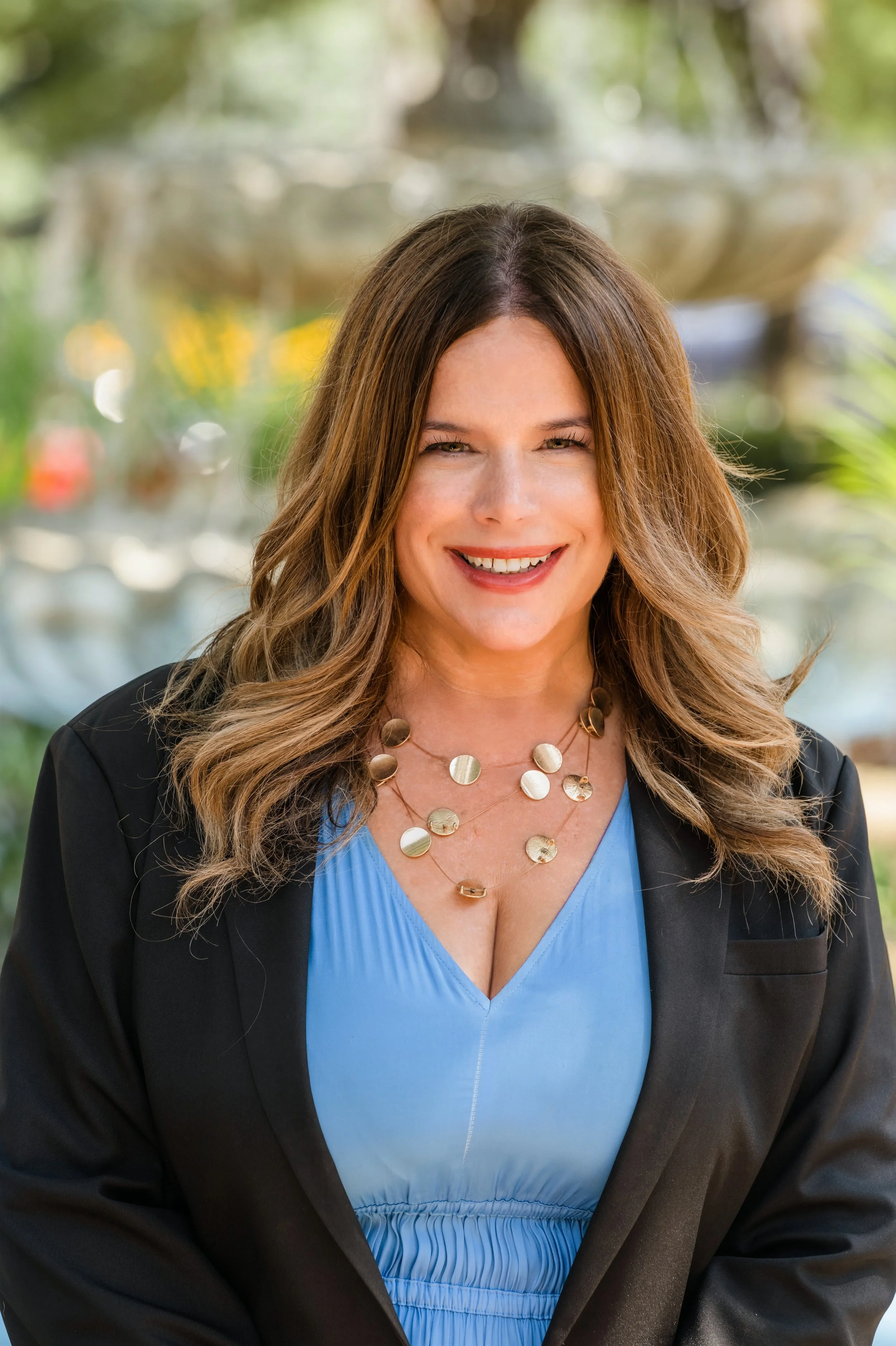 A woman with long, wavy brown hair, wearing a black blazer over a blue dress with a pleated neckline, and a layered gold necklace, smiling outdoors with a blurred fountain and greenery in the background.