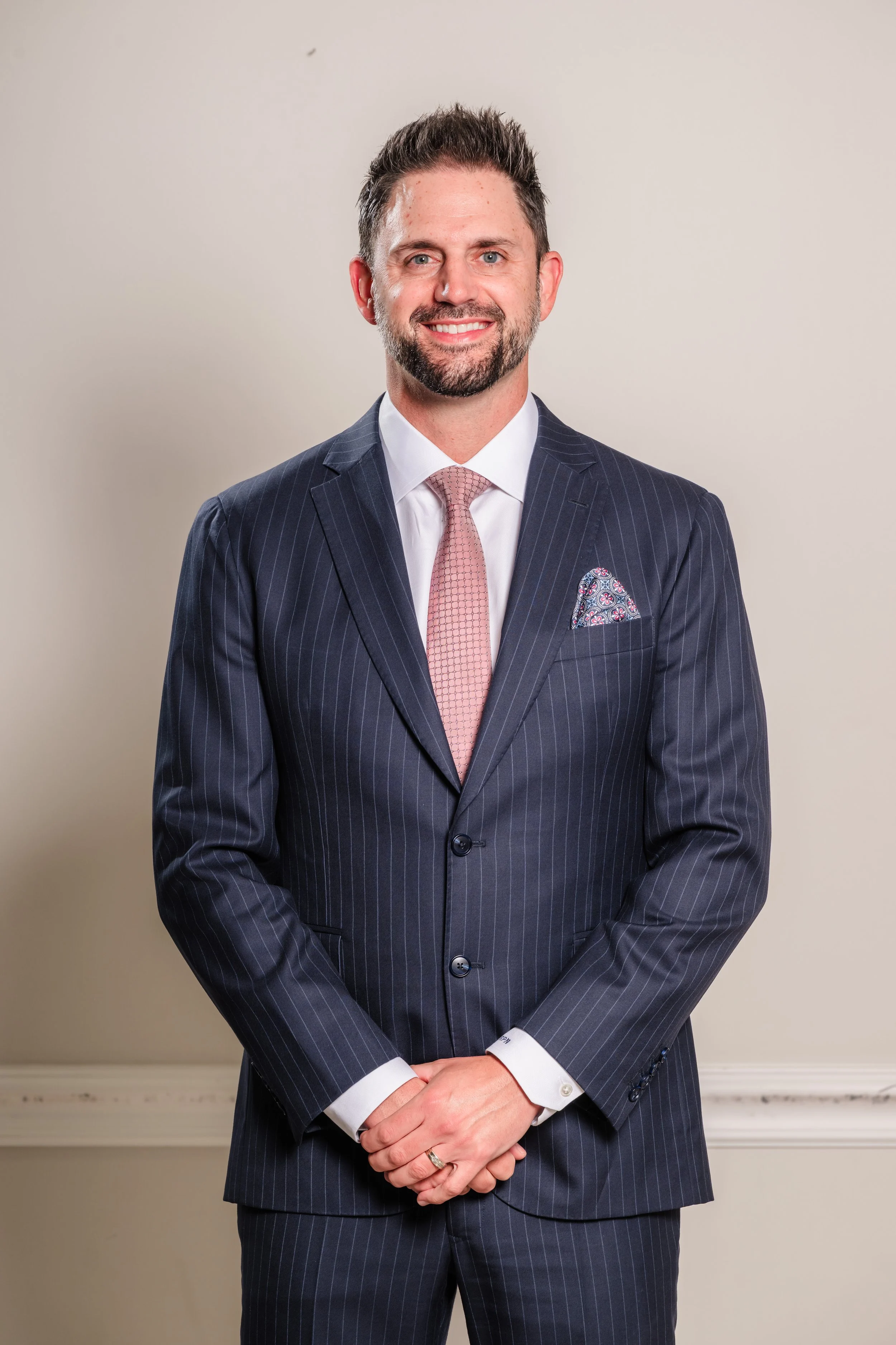 A man in a dark pinstripe suit with a white shirt, pink tie, and patterned pocket square standing against a plain wall.