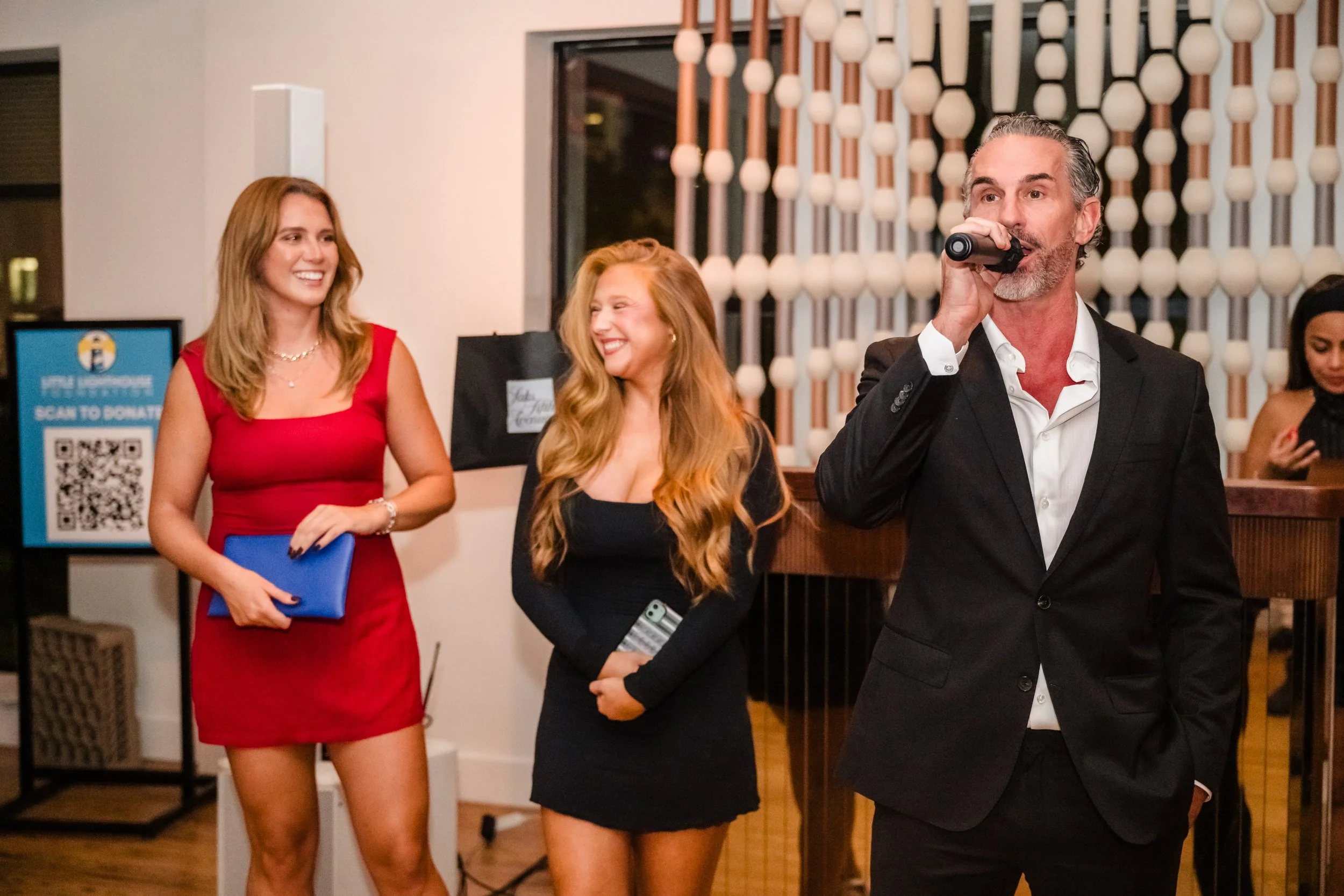 Man in a black suit speaking into a microphone at an indoor event, with two women in dresses listening and smiling in the background.