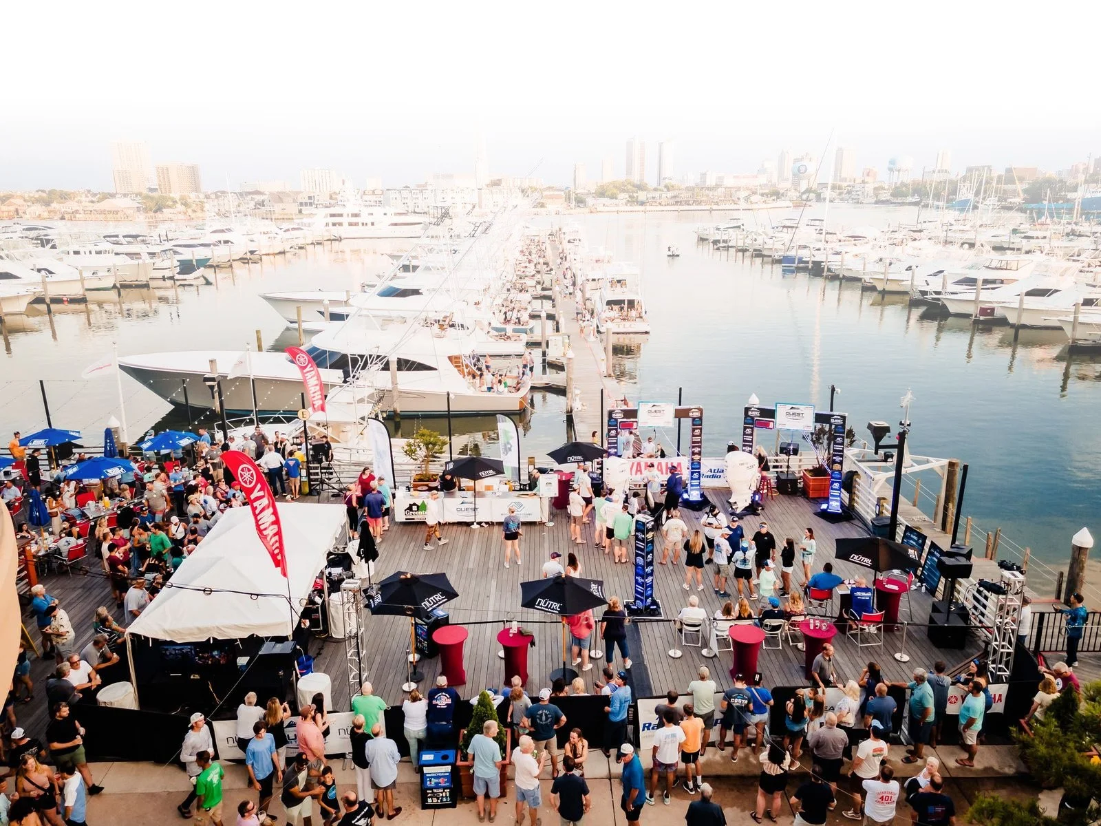 People gathered on a dockside at a boat marina during an event, with boats and yachts docked in the background.