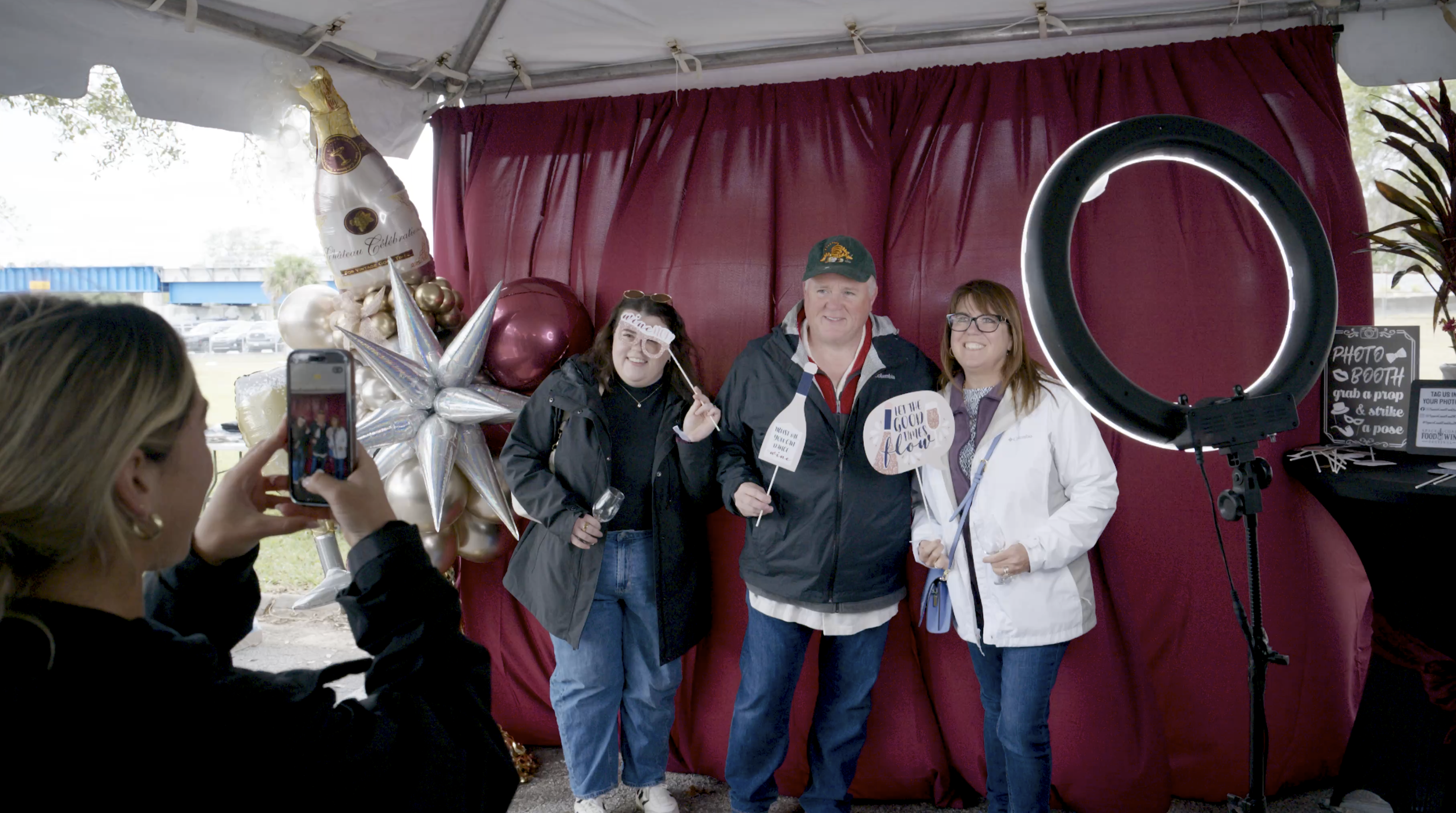 Three guests holding wine-themed photo props posing in front of a red backdrop with balloon decorations at the Space Coast Food & Wine Festival photo booth in Melbourne, Florida