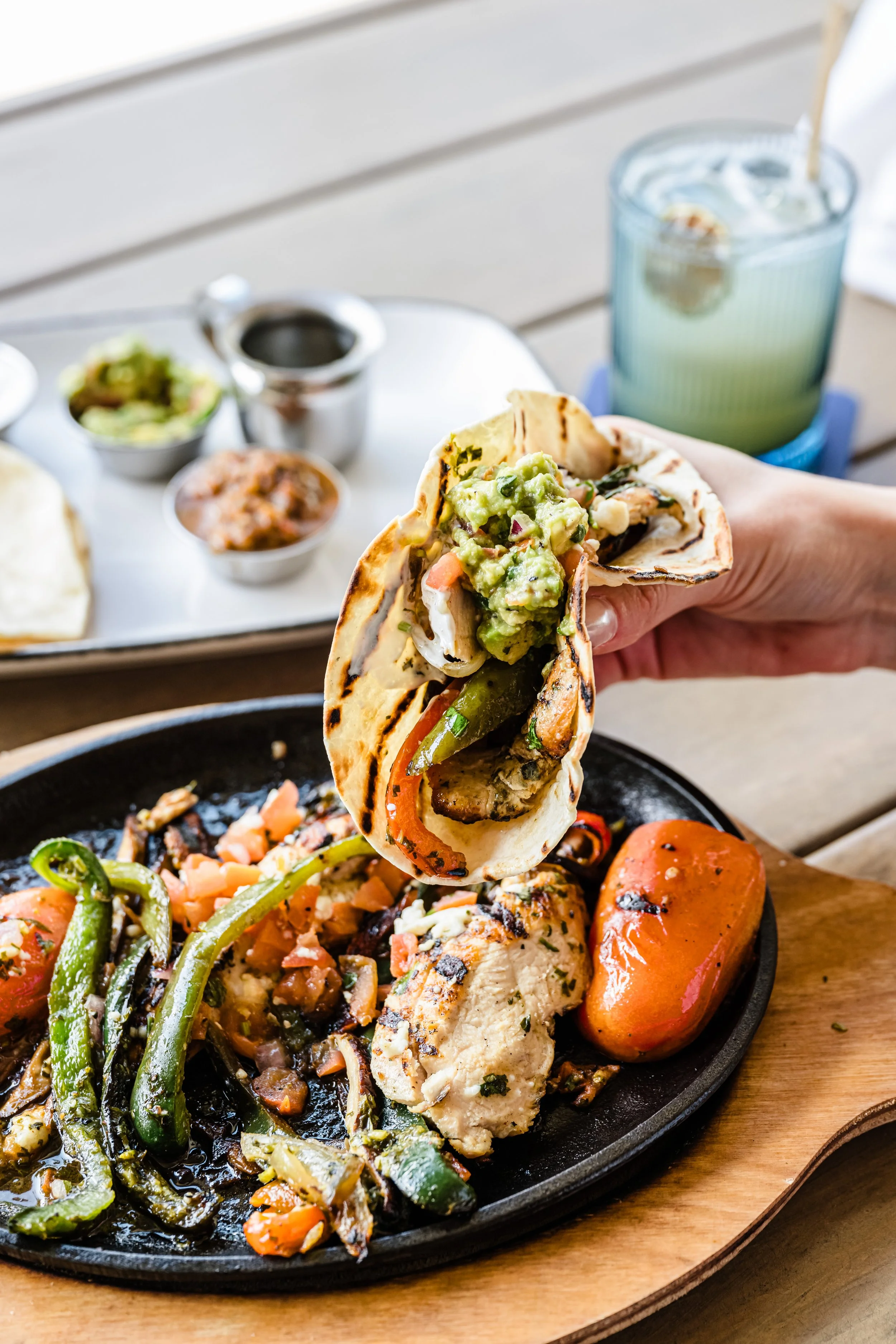 Close-up of a hand holding a grilled chicken taco with guacamole on top, above a sizzling cast iron plate with grilled vegetables, chicken, and a side of roasted tomato, with a beverage and salsa on a white tray in the background.