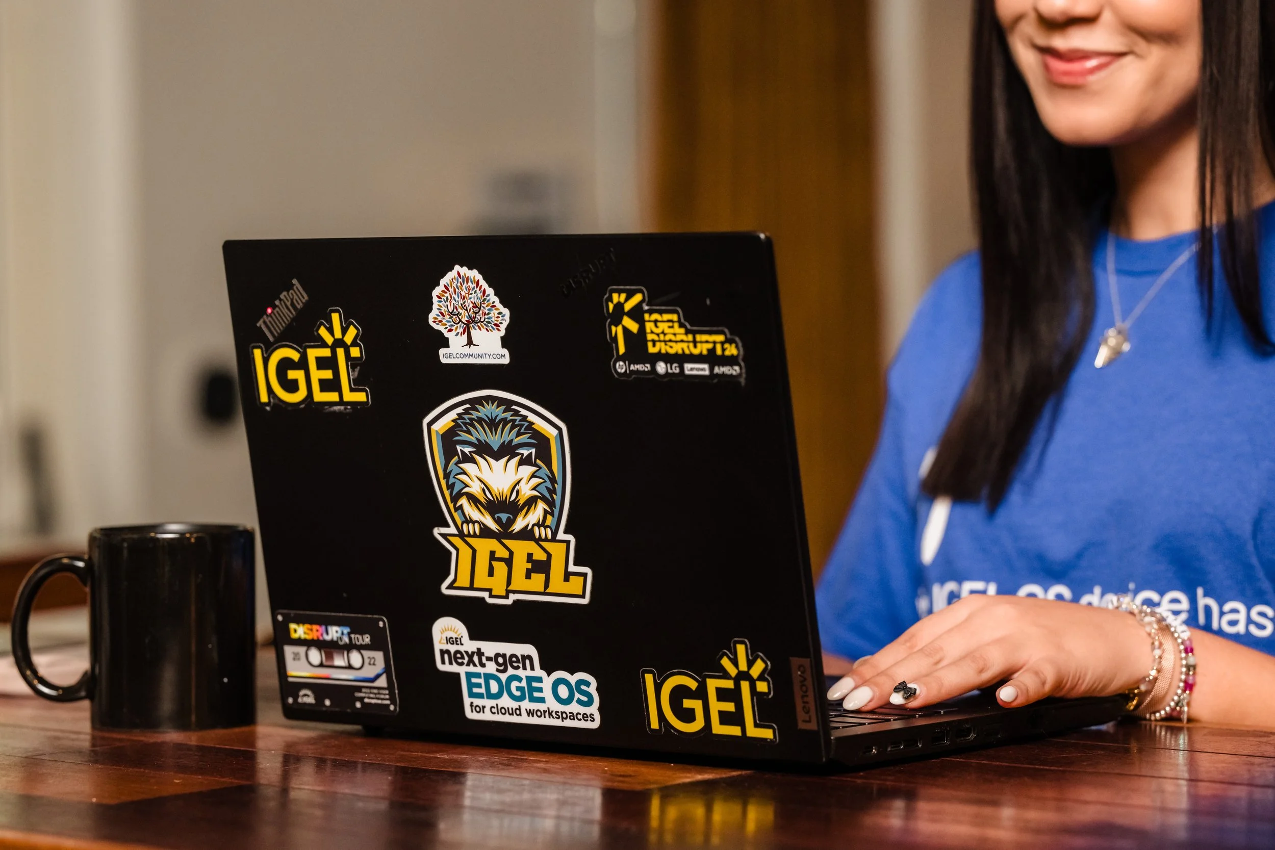 A woman sitting at a wooden table using a black laptop covered with stickers, including Igel, Edge OS, and Disrupt Tour, beside a black mug.