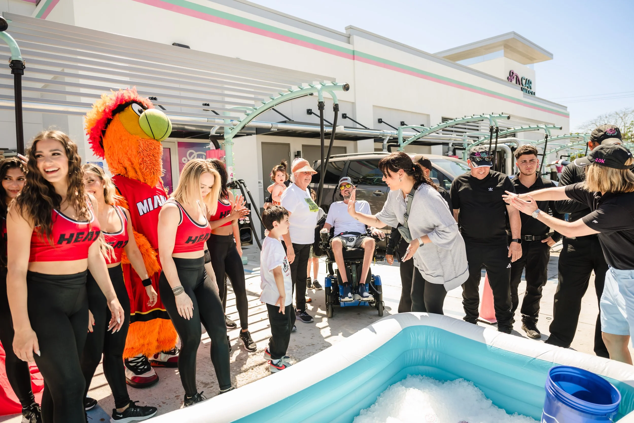 Group of cheerleaders and people gathered around a small inflatable pool filled with ice, outside of a building, with a mascot dressed as a lion and several others cheering and taking photos.