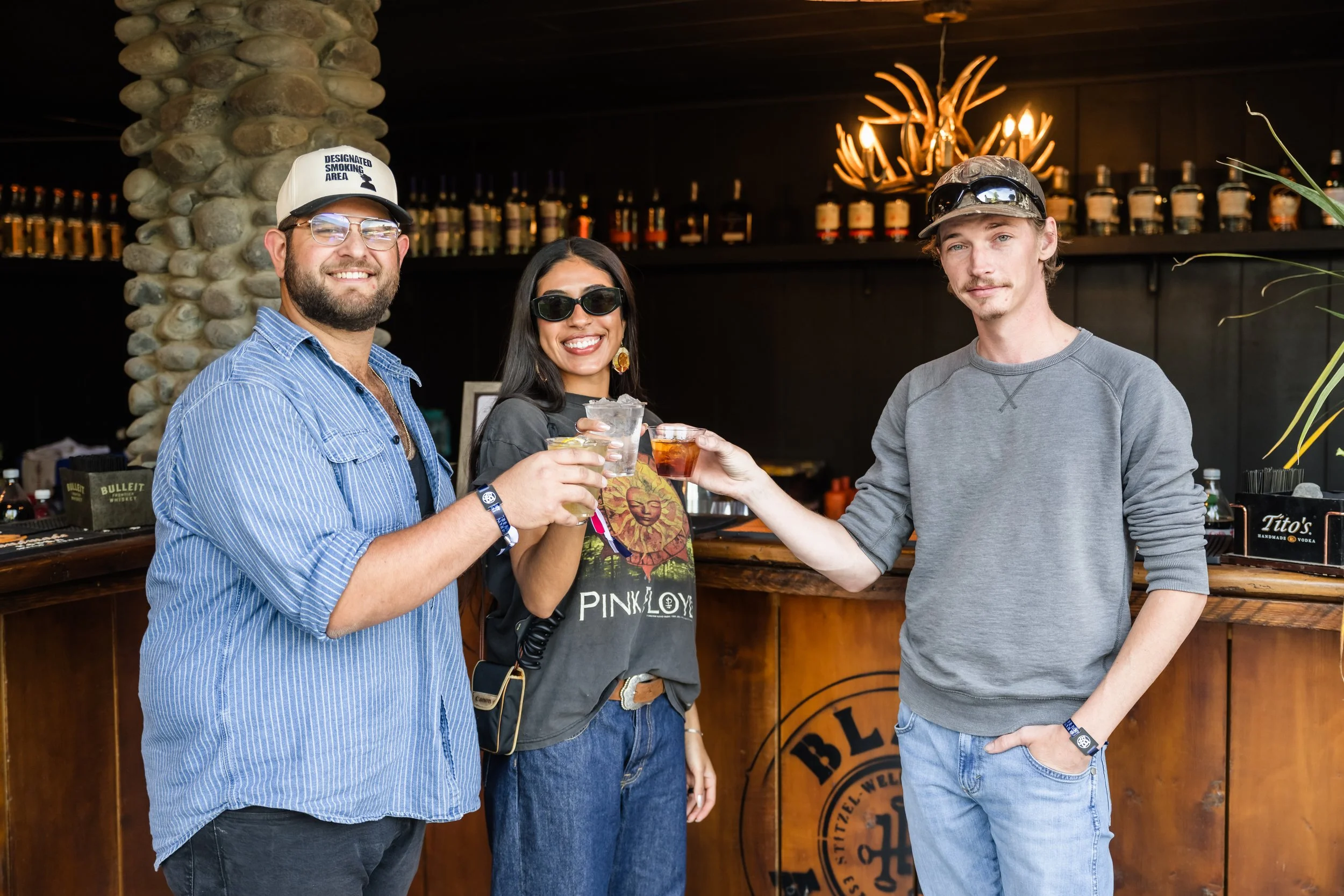 Three people at a bar celebrating with drinks, smiling and toasting each other. The bar has a rustic decor with bottles on shelves and an antler chandelier.
