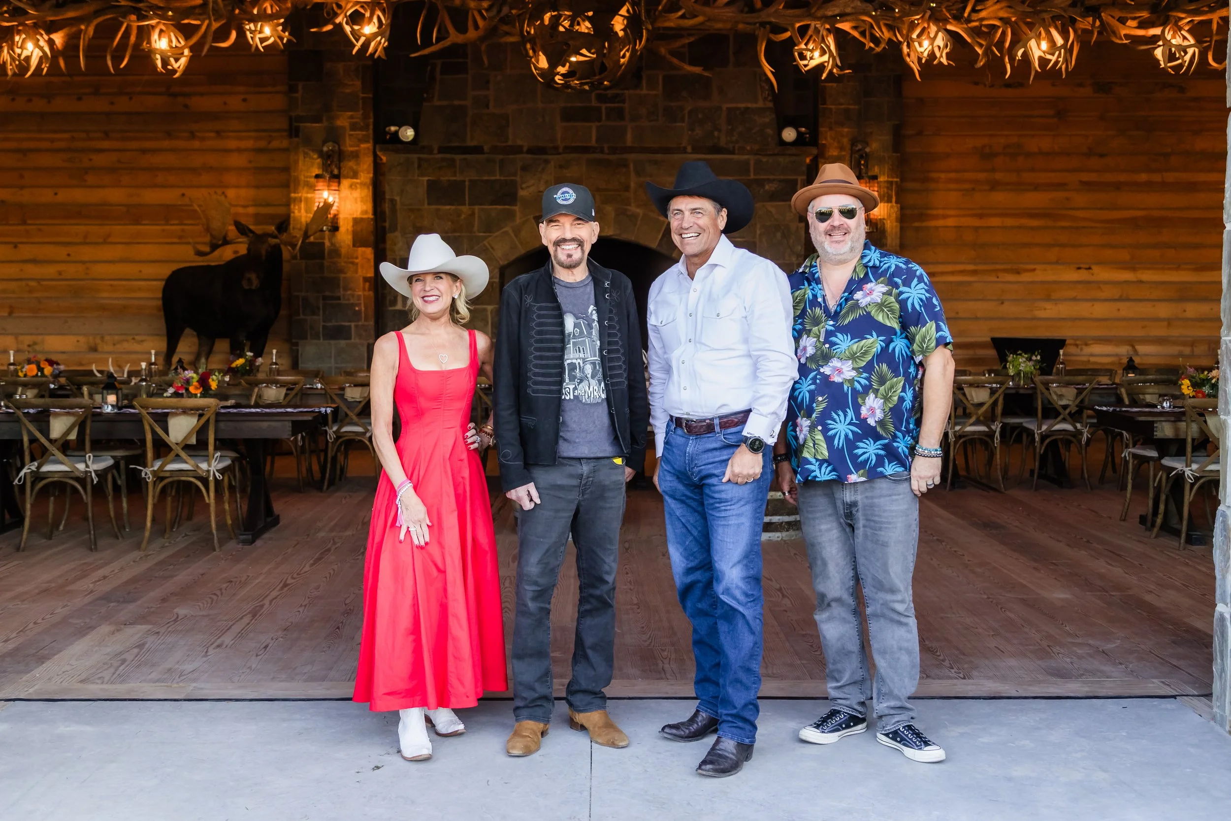 Four people standing inside a rustic, wood-paneled room with a mounted deer head and a large stone fireplace in the background. The people are dressed in casual and country-themed clothing, smiling for the camera.