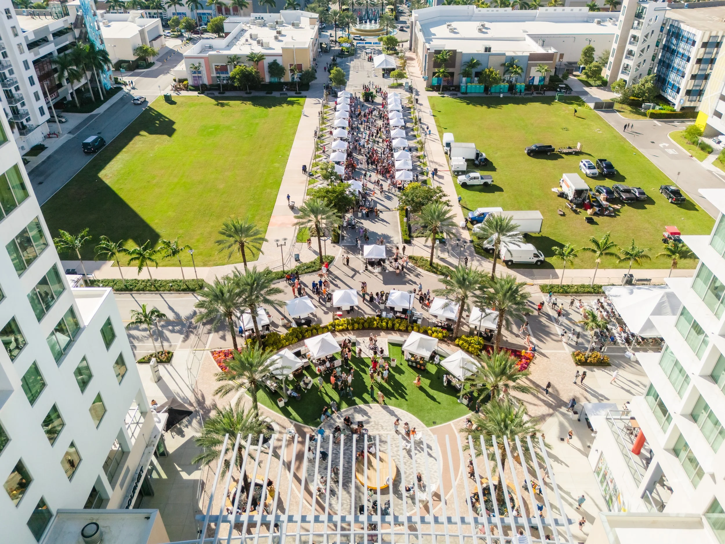 An aerial view of a city square with tents and crowds, surrounded by modern white buildings, green lawns, and palm trees.