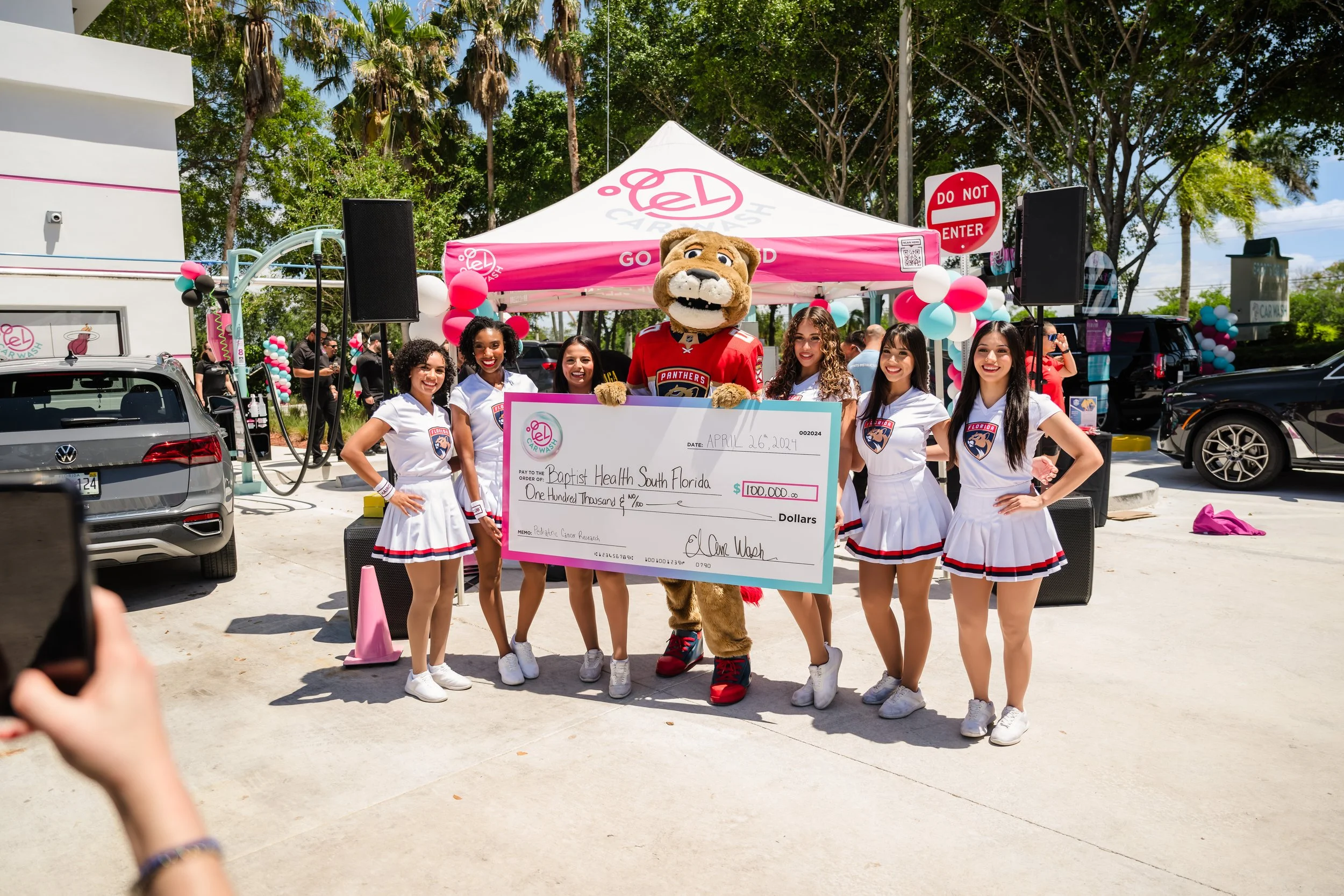 Group of cheerleaders and a person in a panther mascot costume holding a large check for $100,000 in front of a pink and white tent decorated with balloons, during a charity event outdoors.