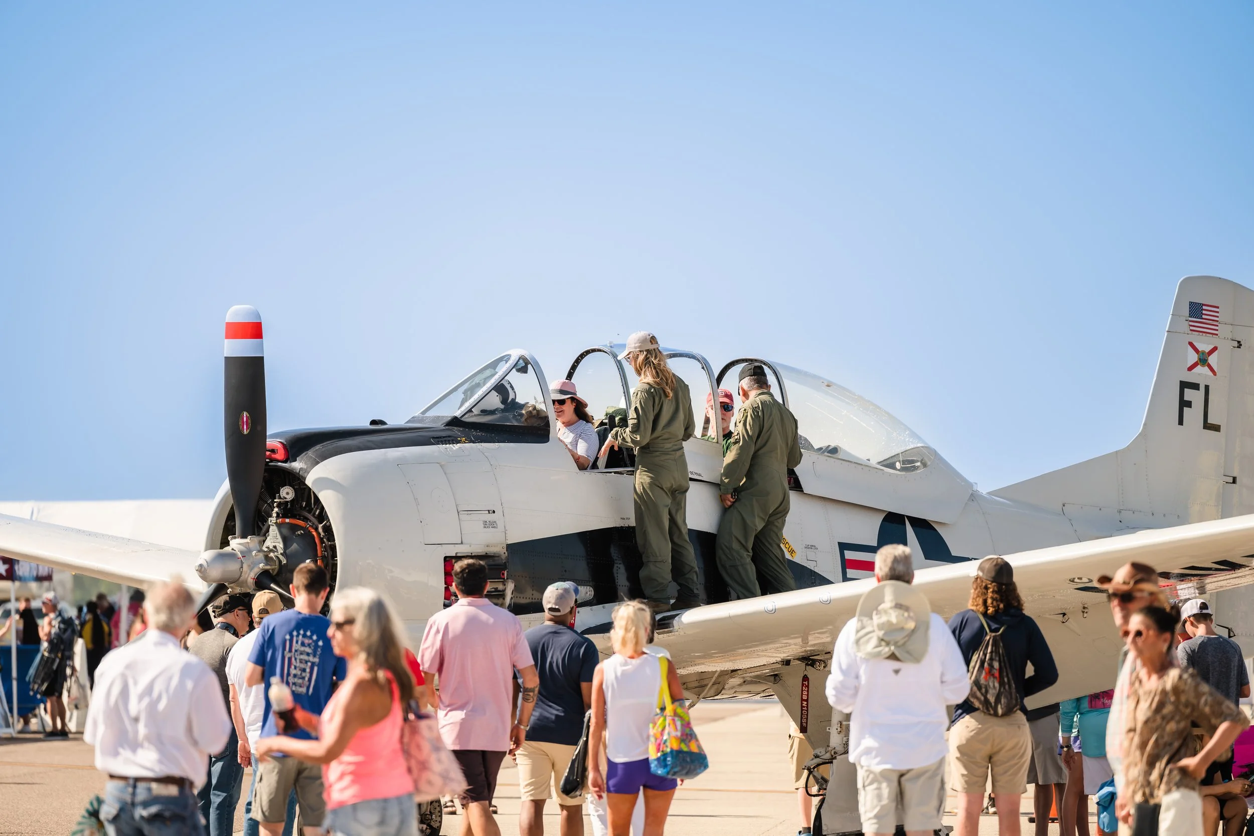 People gathered around a military jet aircraft at an airshow, with some crew members inside the cockpit and others observing, under a clear blue sky.