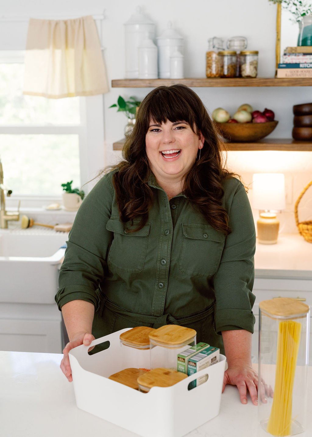 a white woman with long brown hair and bangs wearing an olive green stylish utility suit stands in a white kitchen behind the island holding an organized white bin of glass and wood dry goods containers and pantry staples