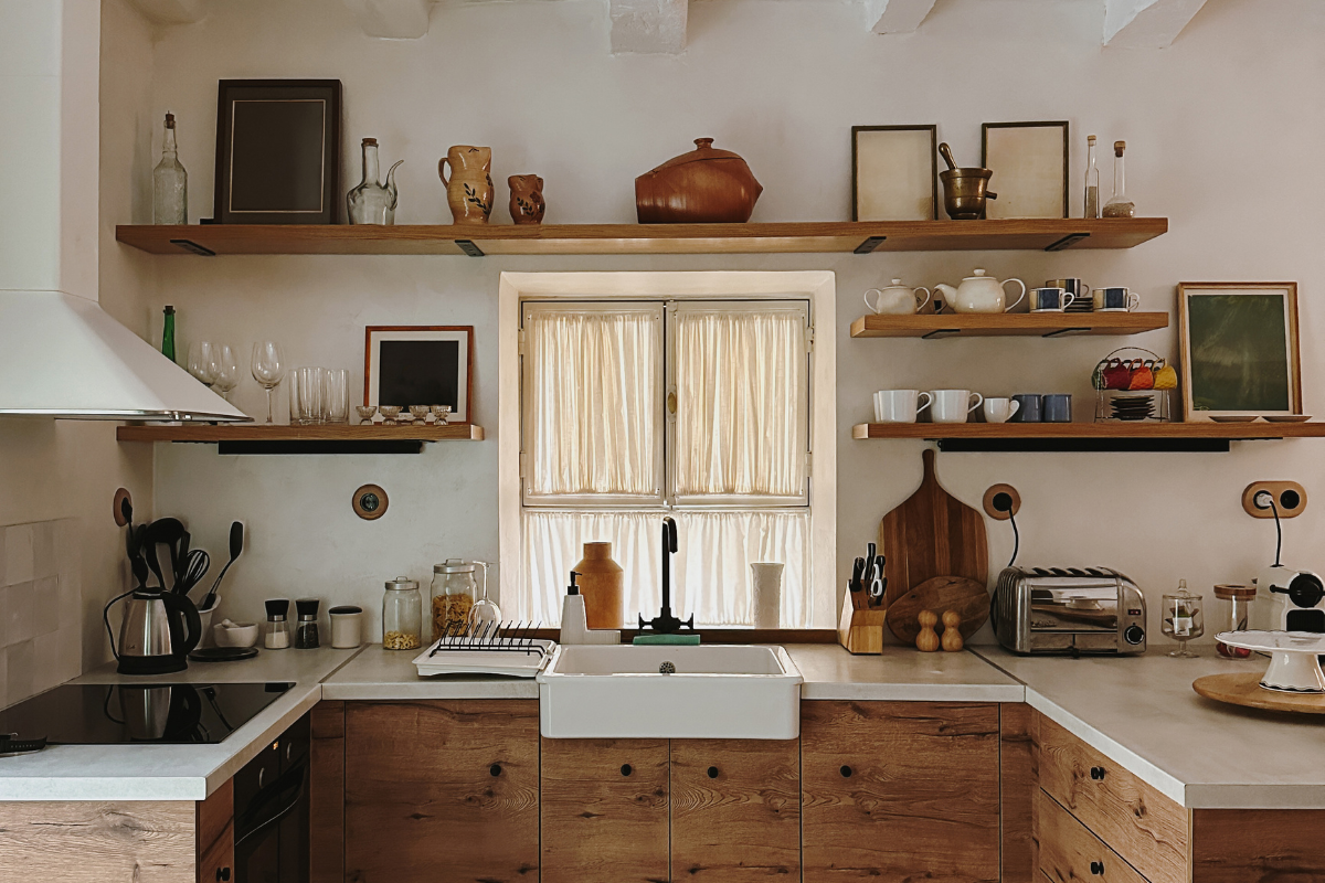 A simple kitchen with neutral cream walls, warmer wood lower cabinets, open upper shelves, a farmhouse sink, and eclectic, aesthetic dishes and decor