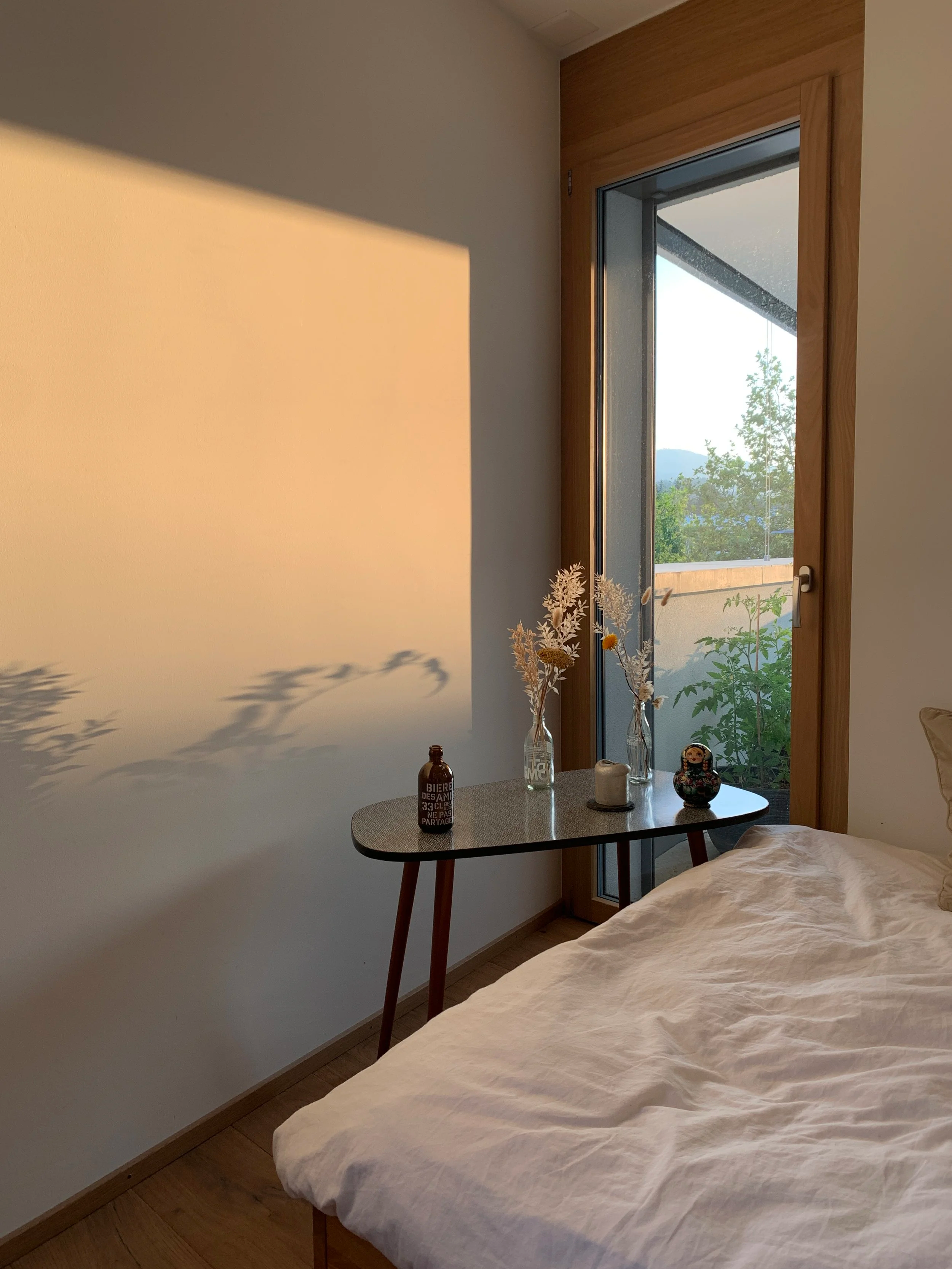 Chambre avec un lit couvert d'une drap blanc, une petite table avec des décorations telles que des vases avec des fleurs, une bouteille et une figurine, une fenêtre laissant entrer la lumière naturelle et offrant une vue sur un paysage avec des arbres et des montagnes.