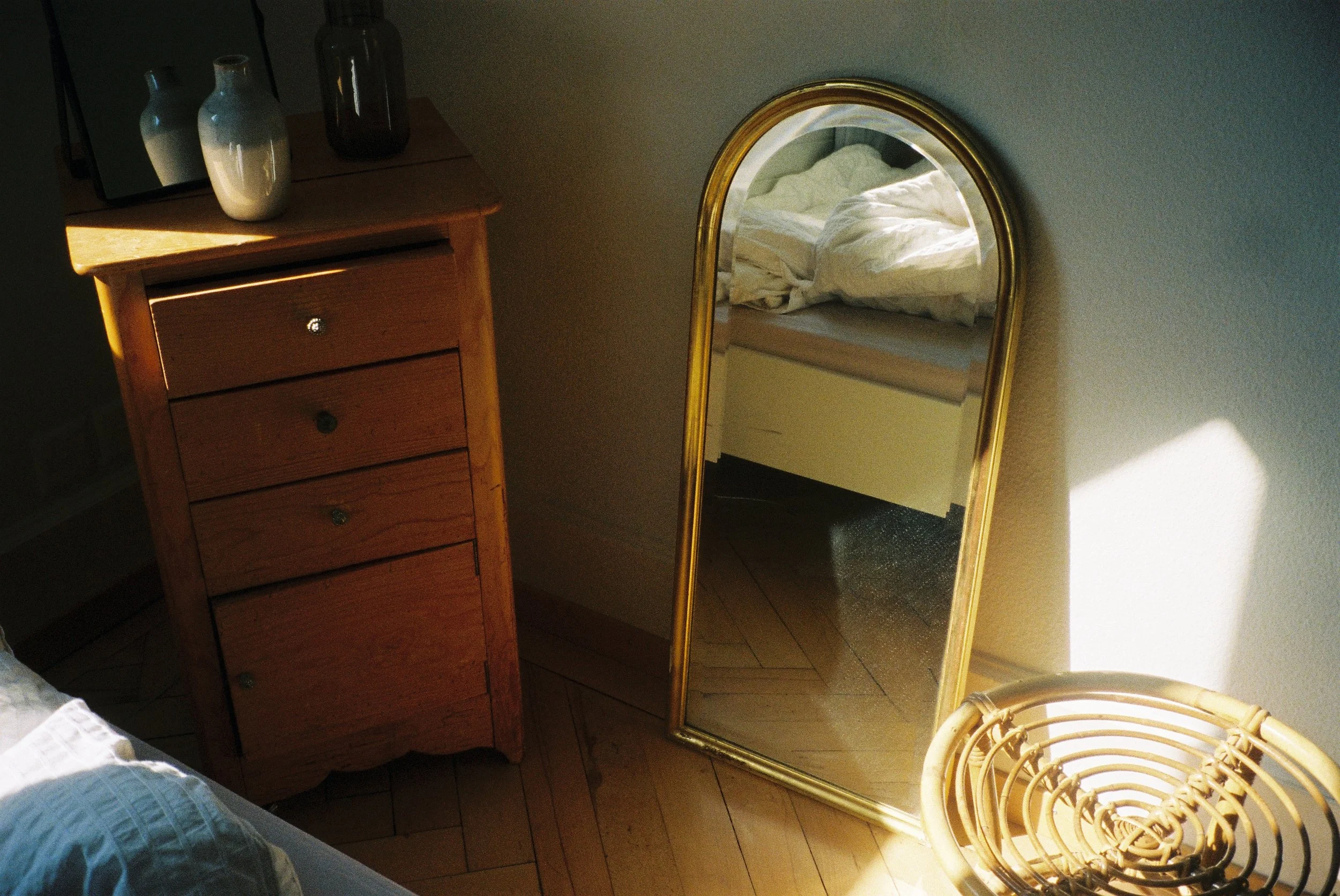 Un miroir ancien avec cadre doré posé contre un mur, reflétant un lit avec draps blancs et une lampe. À côté, une commode en bois avec des vases en céramique et un vase en verre. Une chaise en rotin est partiellement visible.