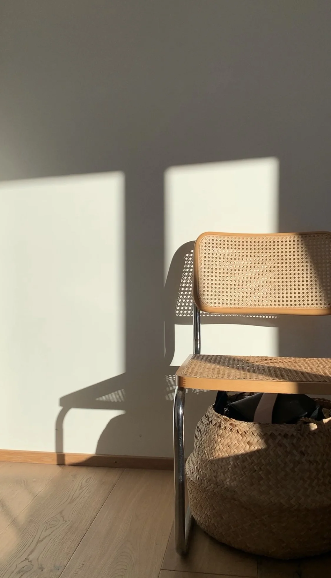 Une chaise en bois avec un dossier en cannage et un cadre en métal est placée contre un mur blanc. L'ombre de la chaise est projetée sur le mur par la lumière naturelle.