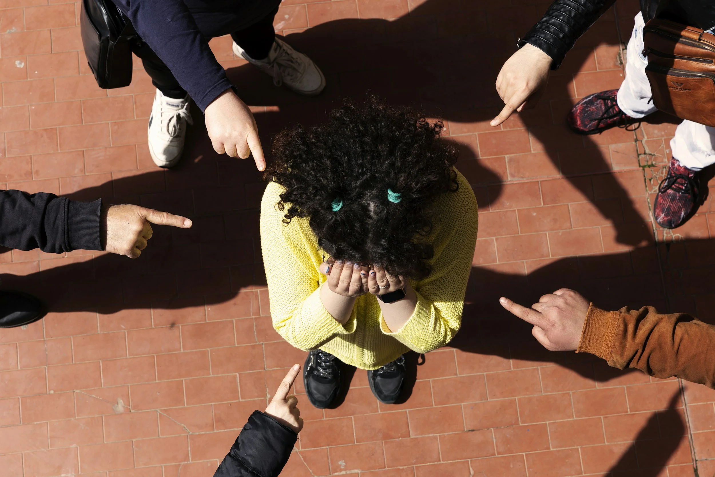 Overhead view of a person sitting on a brick walkway with their face covered while several people point toward them.