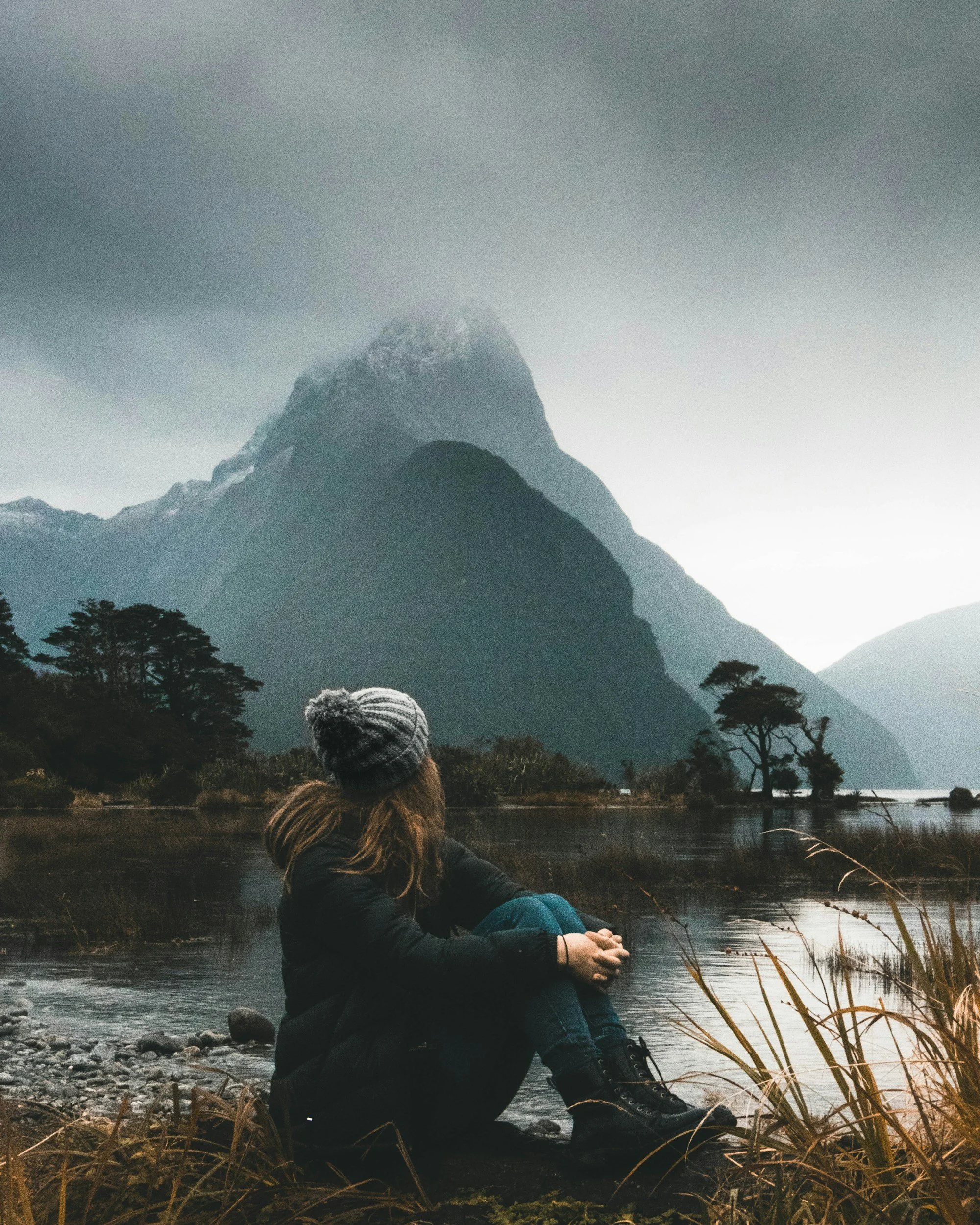 Person sitting beside a lake looking toward a mountain partially covered by clouds.