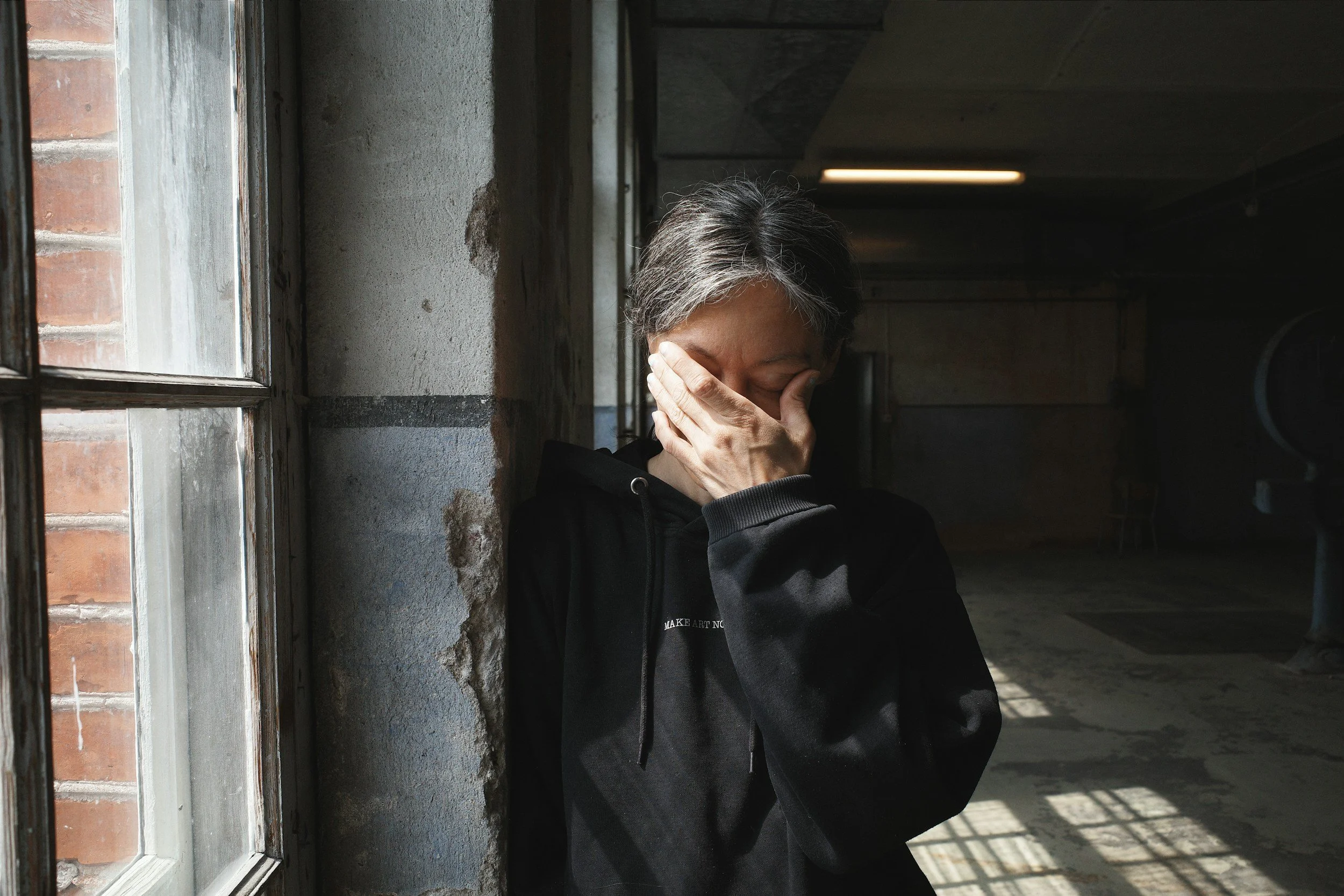 Person standing beside a window with one hand covering their face.