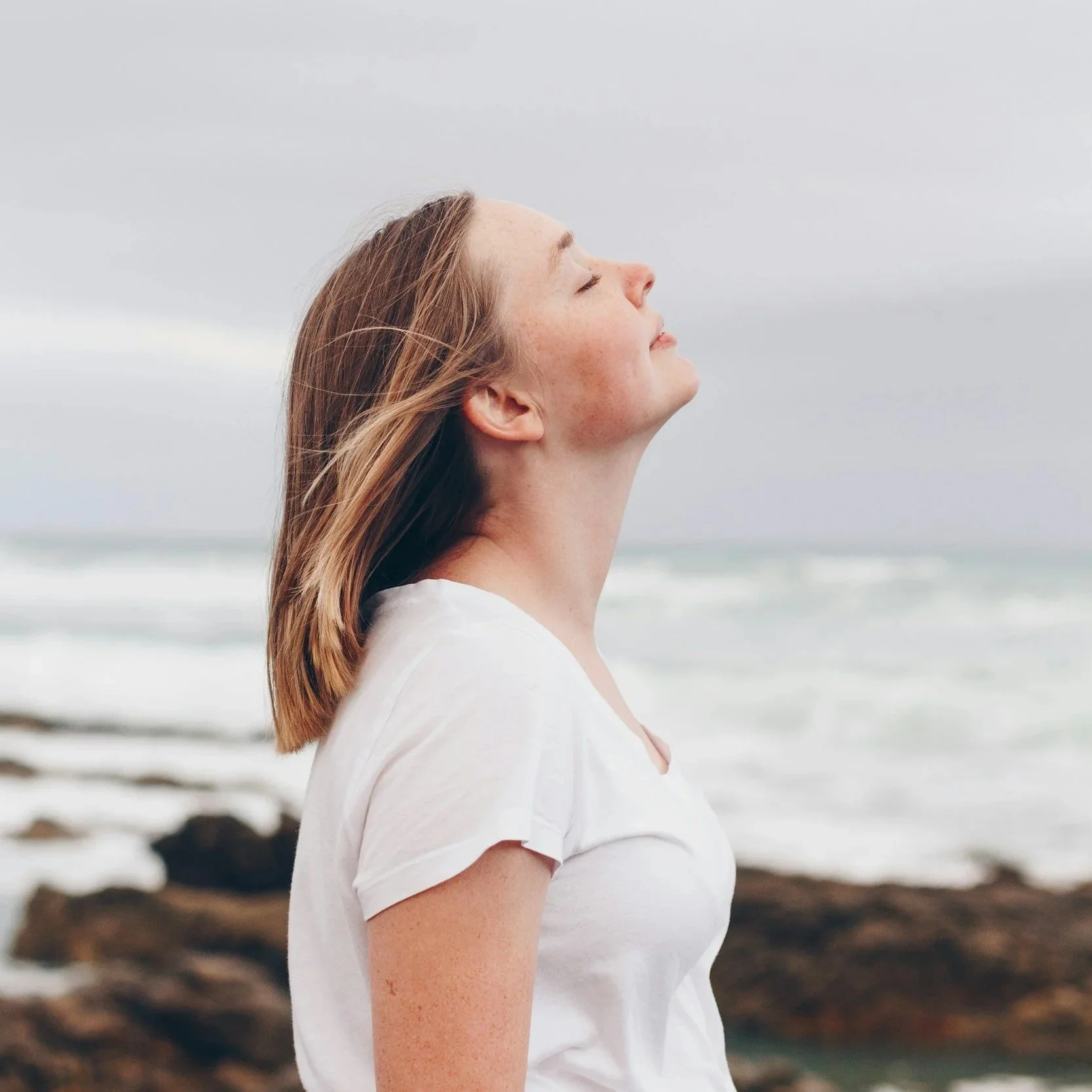 Side profile of a woman standing outdoors near the ocean with her eyes closed.