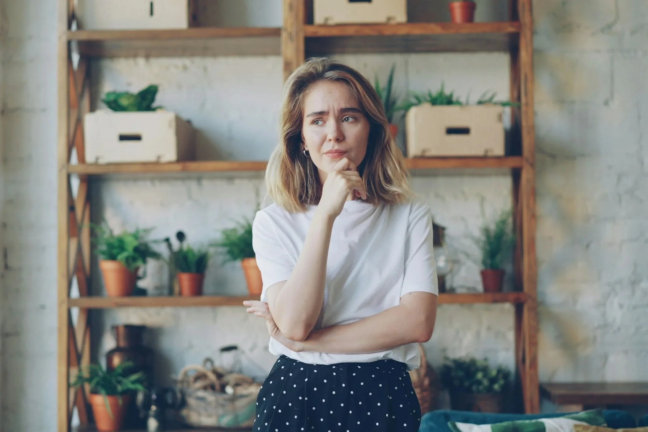 Person standing indoors with one hand on their chin in front of wooden shelves with potted plants.